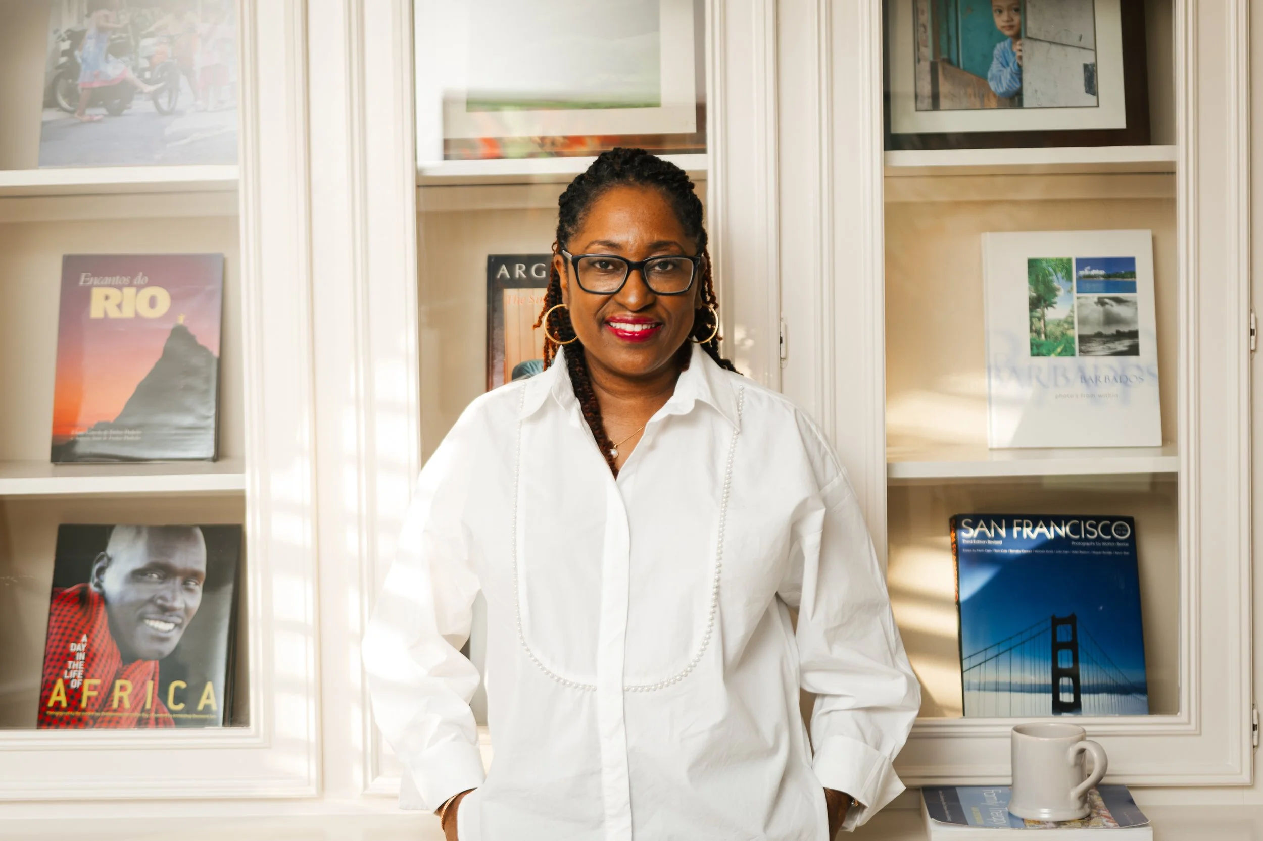 A woman with glasses and earrings wearing a white shirt, standing in front of a bookshelf with travel books and photos, and holding her hands in her pockets.