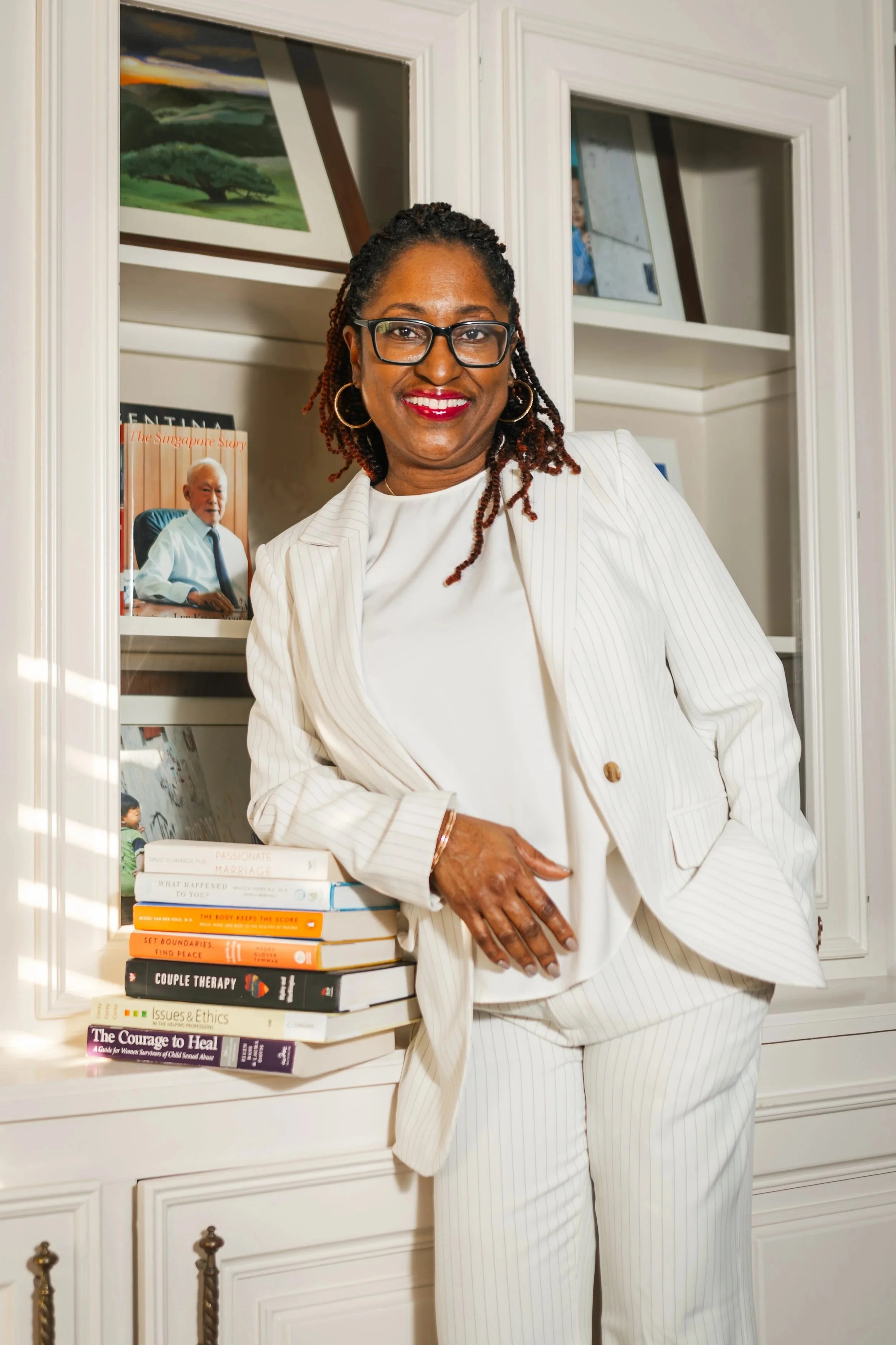 A woman in a white pinstripe suit standing next to a stack of books on a white cabinet, smiling at the camera, with a bookshelf and framed pictures in the background.