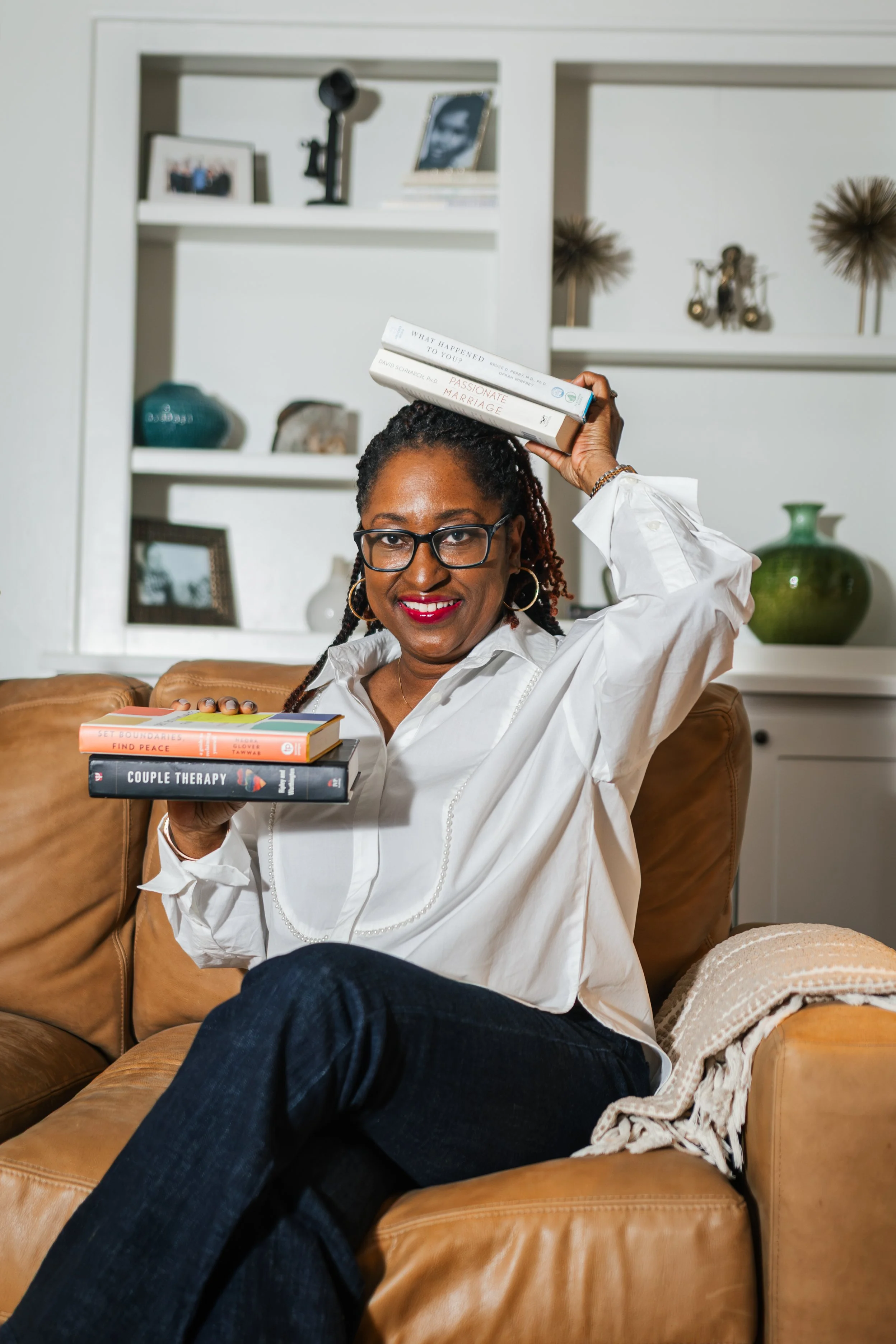 A woman with glasses and earrings sitting on a leather couch, holding books in her lap and resting a book on her head, in a cozy living room with decorative items on shelves behind her.