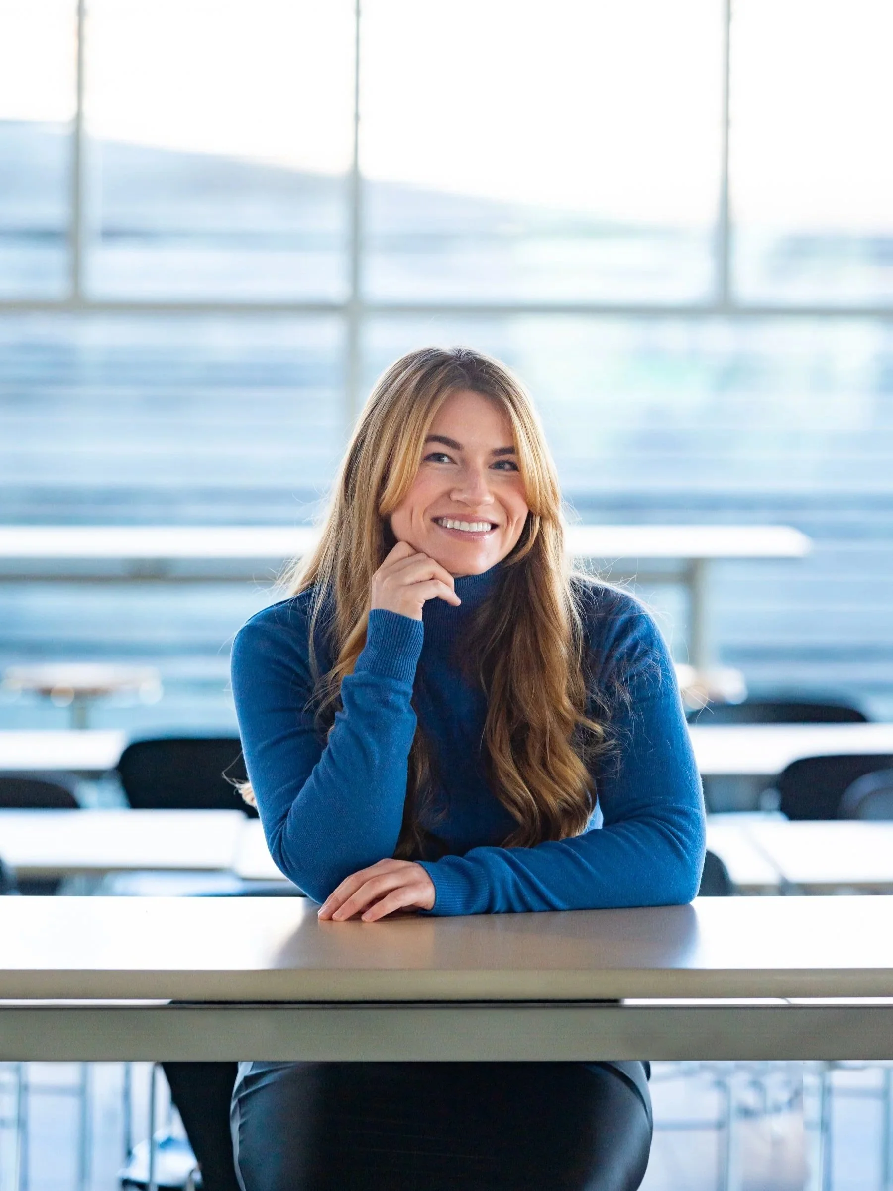 A photo of Kelly Firth, a smiling woman with long hair in a blue sweater sitting at a desk in a bright office with large windows.