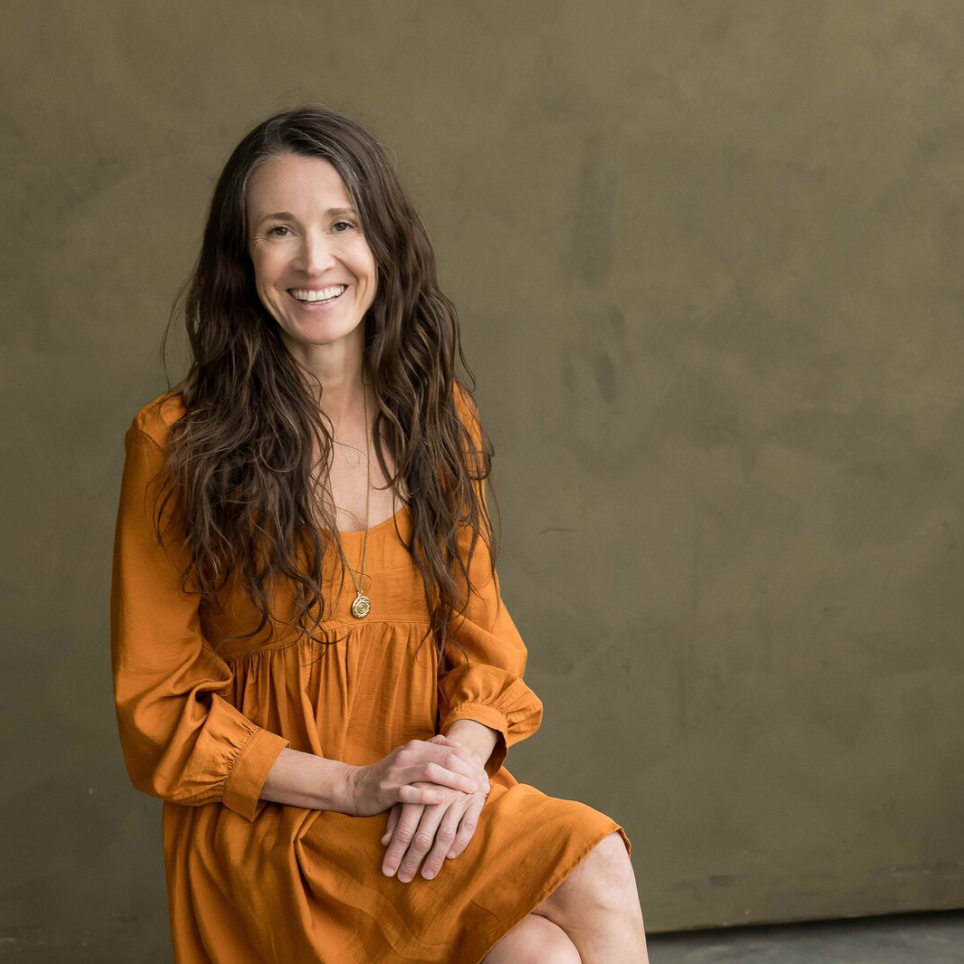 A woman with long, wavy brown hair is sitting and smiling at the camera, wearing an orange dress and a pendant necklace, against a plain, muted background.
