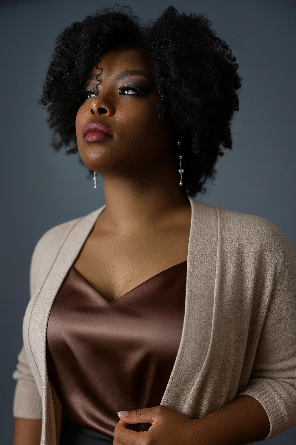 A portrait of a Black woman with curly hair wearing earrings, a beige cardigan, and a brown top, looking slightly upwards against a gray background.