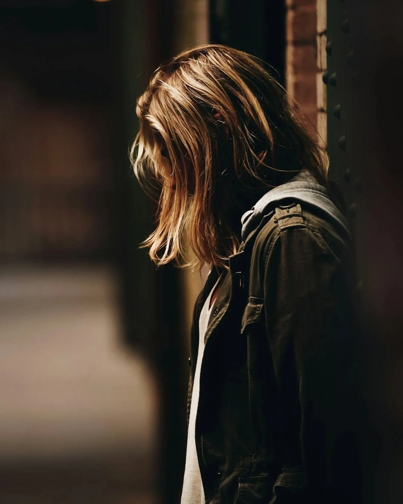 A woman with long brown hair, wearing a dark jacket and a backpack, standing alone in a dimly lit area, leaning against a brick wall with her head down.