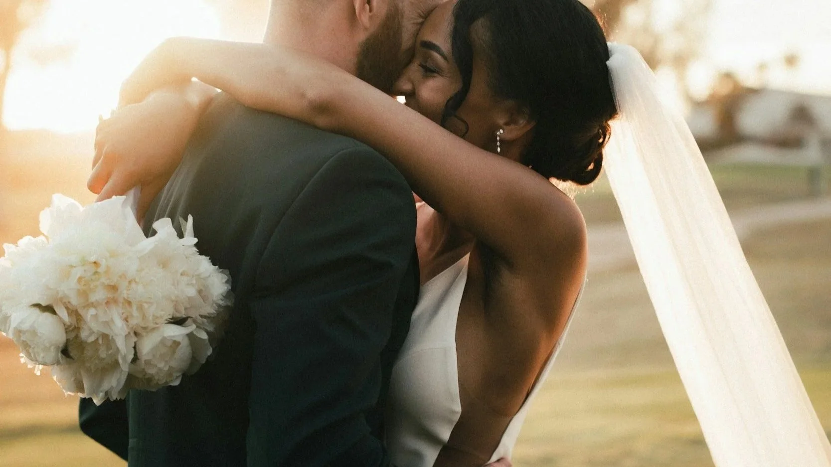 A newlywed couple sharing a close dance or embrace outdoors during sunset, with the bride wearing a white wedding dress and veil, and the groom in a dark suit. The bride is smiling and holding a bouquet of white flowers.