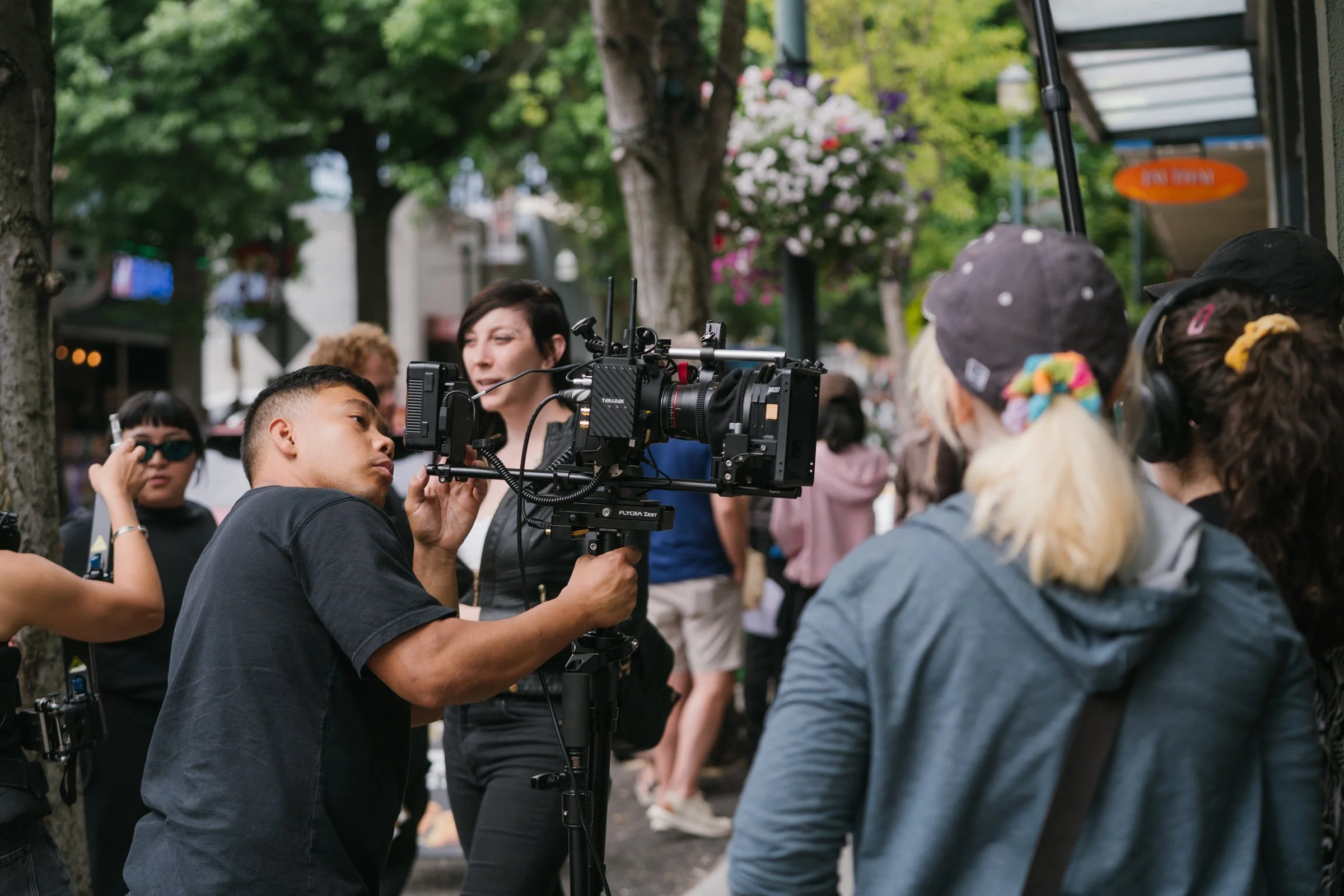Filmmaking crew setting up camera shot on a busy outdoor city street with trees and hanging flower baskets.