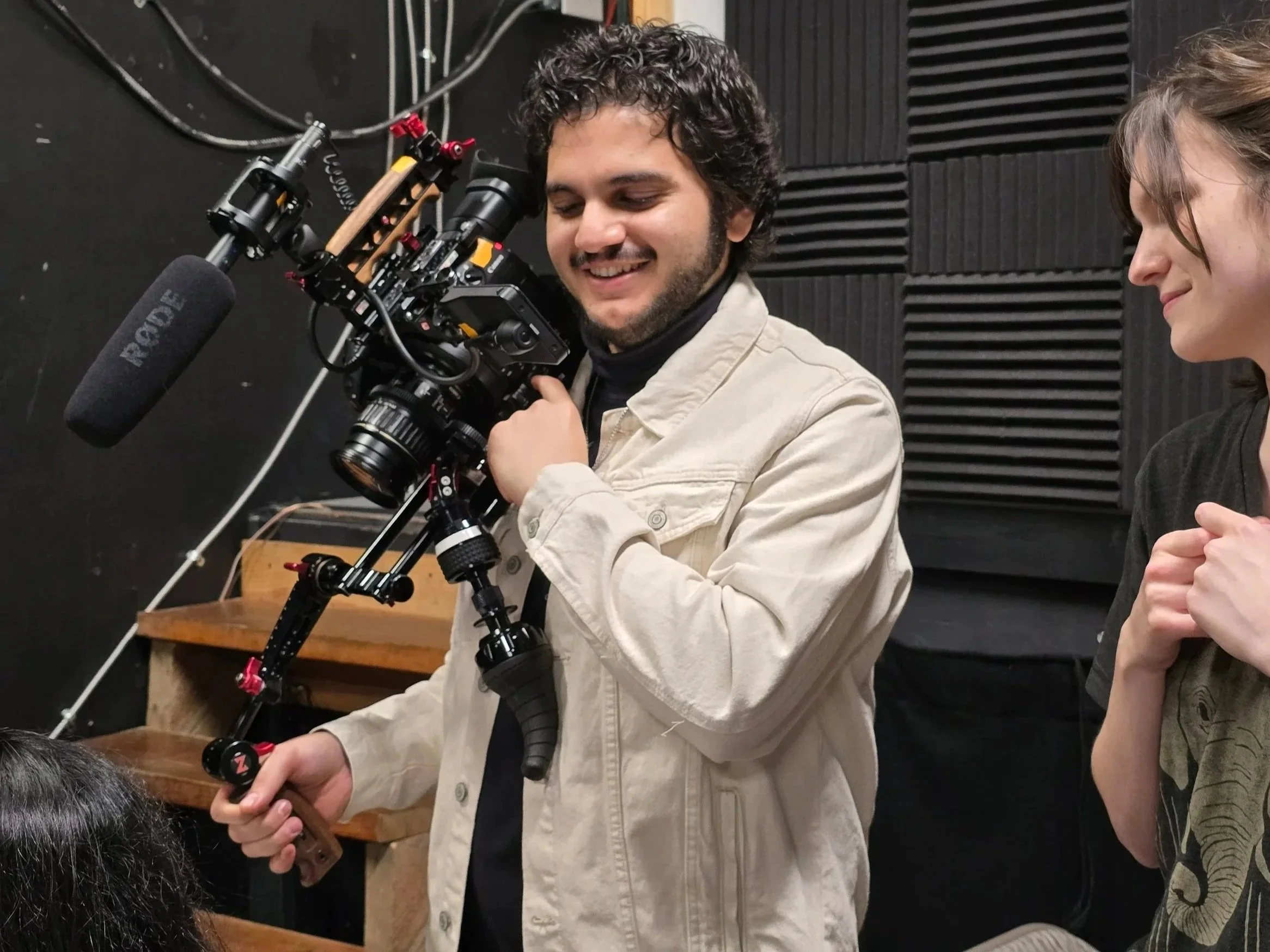 A man operating a professional camera with a microphone attached, while smiling, and a woman standing beside him, both indoors with soundproof foam on the walls.