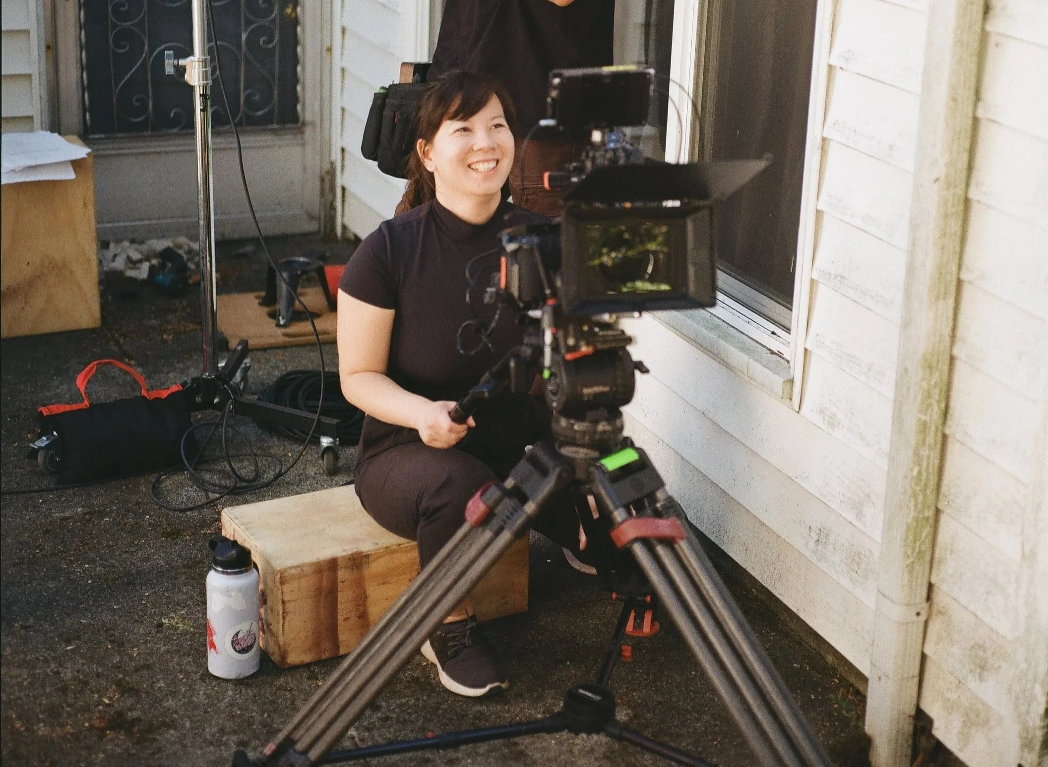 A woman with short dark hair, wearing a black shirt, sitting on a wooden block beside a white wooden shed, smiling while operating a professional video camera on a tripod for filming.