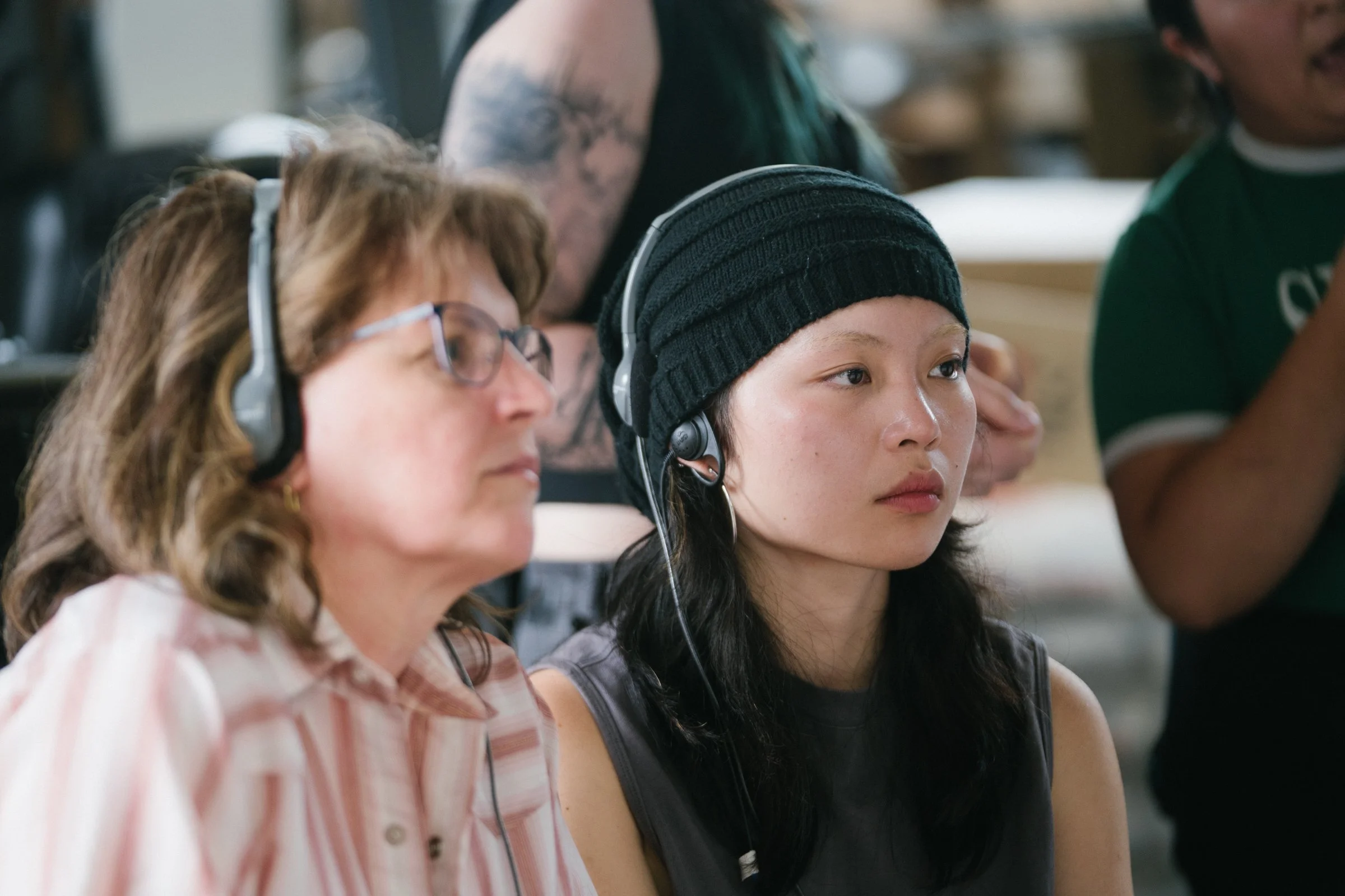 Two women wearing headphones attending a listening session, one older woman with glasses and a striped shirt, and a young woman with a black beanie and sleeveless top, in a room with other people and books in the background.