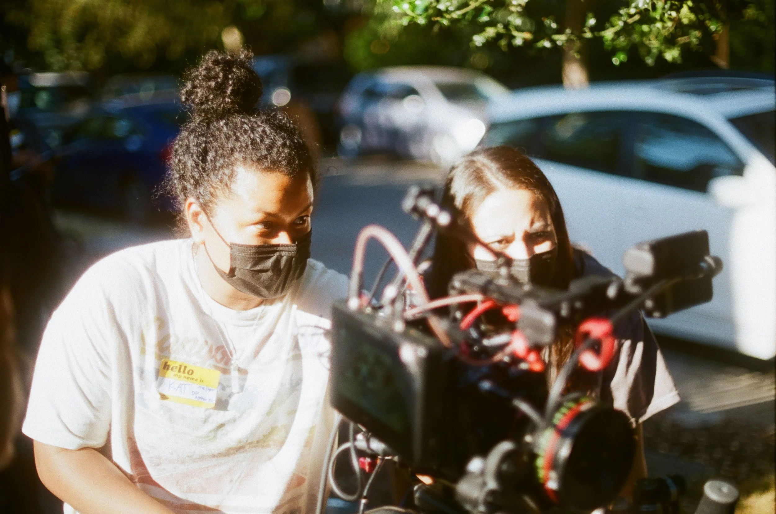 Two women wearing face masks look at a professional film or camera equipment outdoors in a parking lot with parked cars and trees in the background.