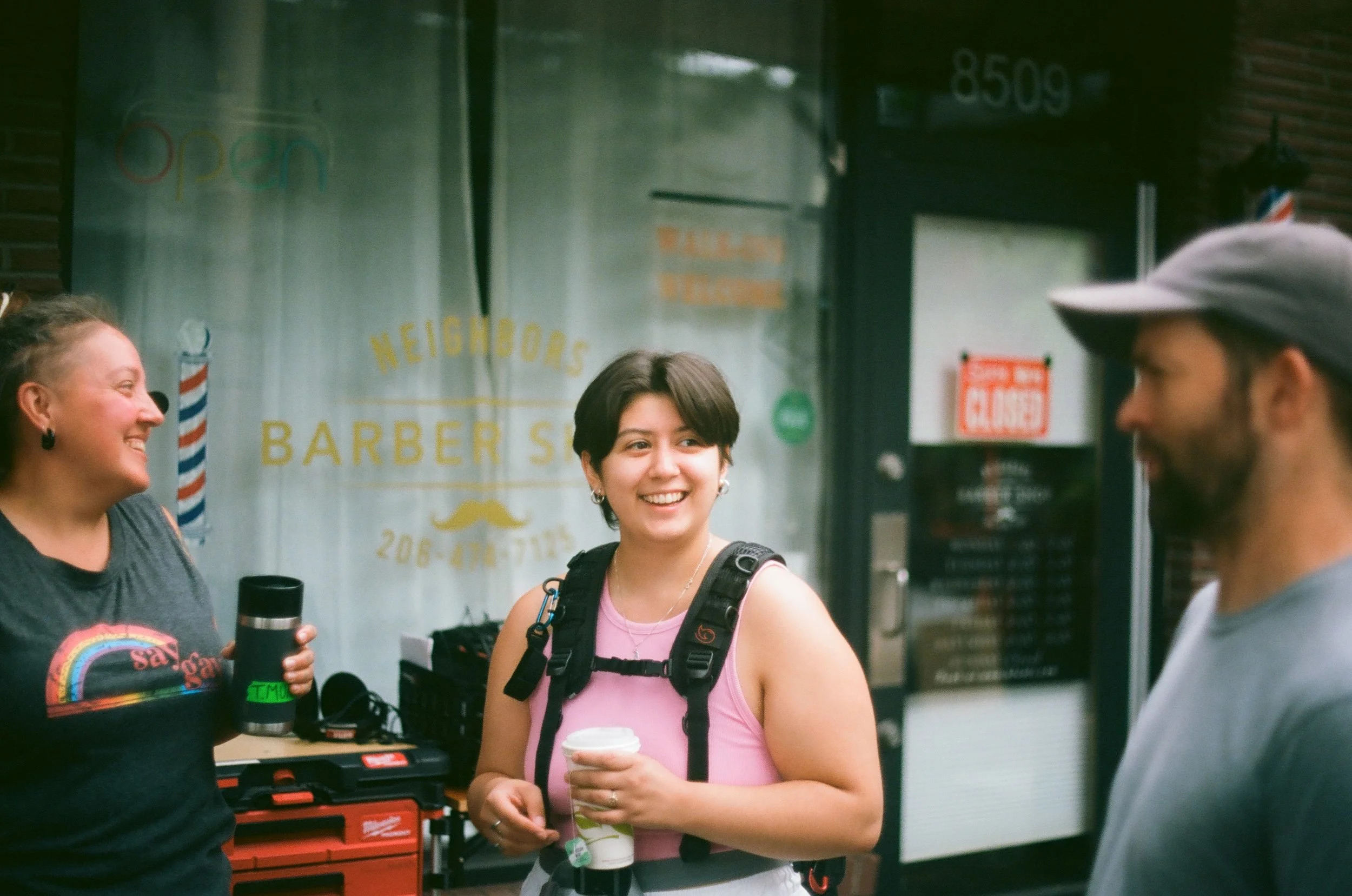 Three people are standing and smiling outside a storefront with a sign that reads 'Neighbors Barber Shop.' One woman in the center is holding a coffee cup and wearing a backpack. The woman on the left is holding a thermos, and the man on the right is wearing a cap. The background features barber pole decor and a closed sign.