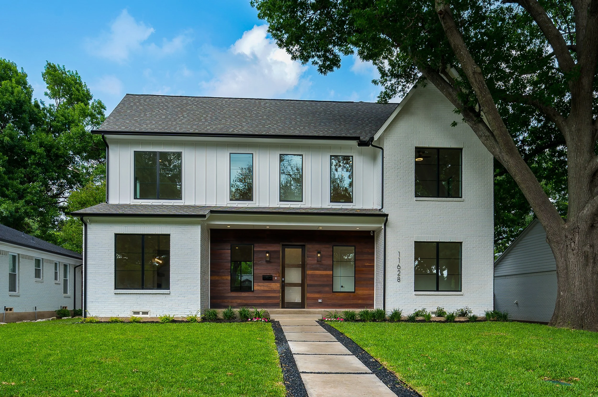 Modern two-story house with white brick and wood exterior, large windows, a front walk, with mature trees in the background located in the Lochwood neighborhood of Dallas, TX
