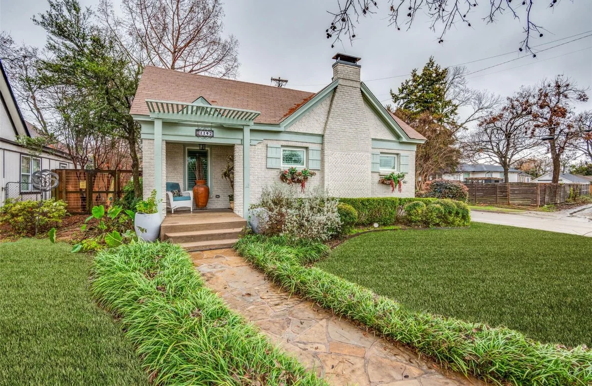 Front yard view of a cozy antique house with a stone pathway leading to the front porch, surrounded by lush green grass and landscaped bushes, trees, and potted plants located in the Elmwood neighborhood of Oak Cliff, Dallas, TX.