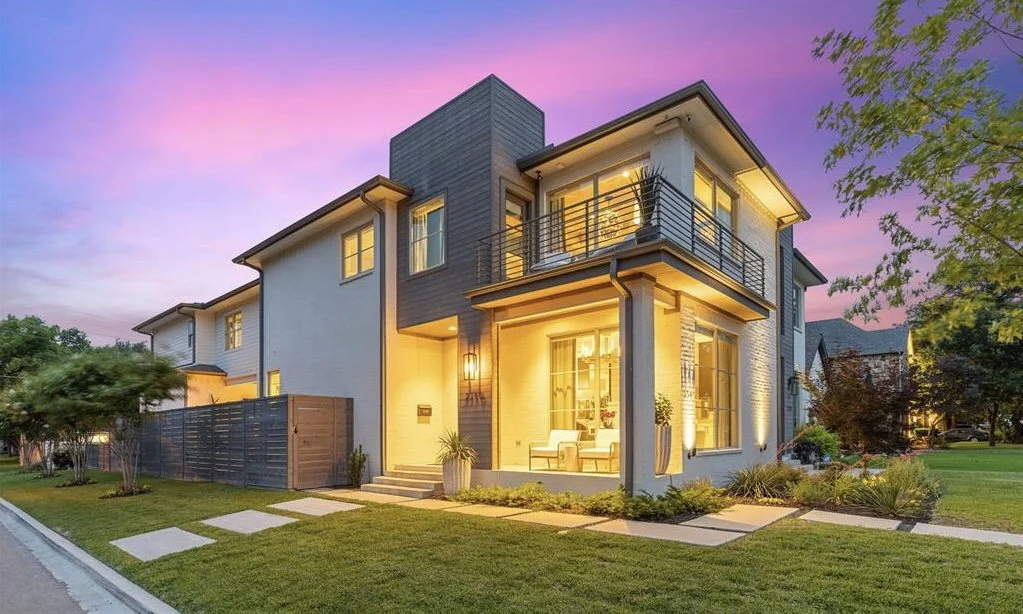 A newer construction modern two-story half duplex built by Newport Homes and designed by Architect Bob Anderson with illuminated windows and a cozy front porch during sunset, surrounded by landscaping in the Glencoe Park neighborhood, Dallas, TX