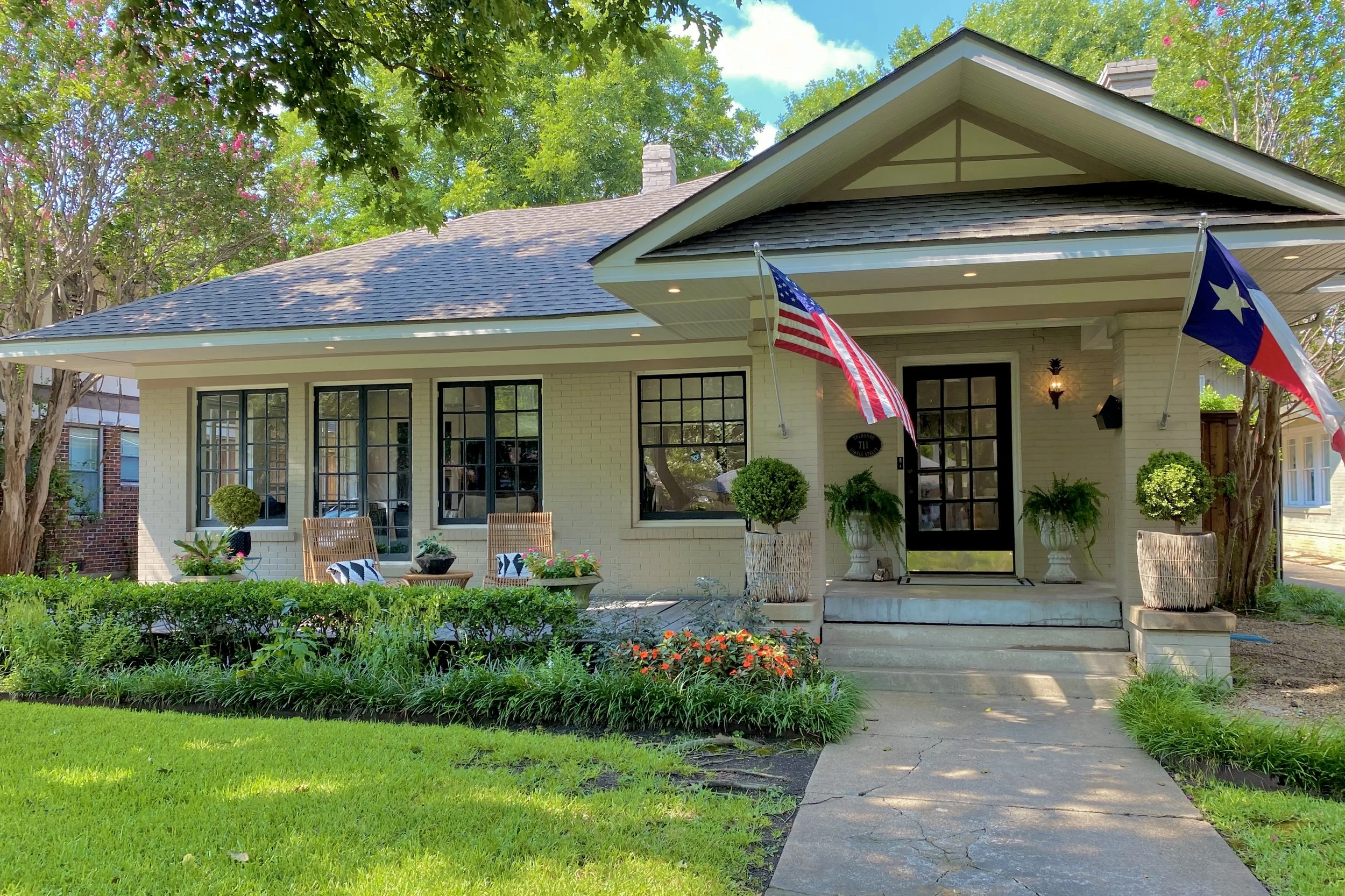 Front view of a white brick Craftsman style house with a porch, two American flags, and a Texas flag, surrounded by green trees and plants in the Junius Heights neighborhood of Dallas, TX.