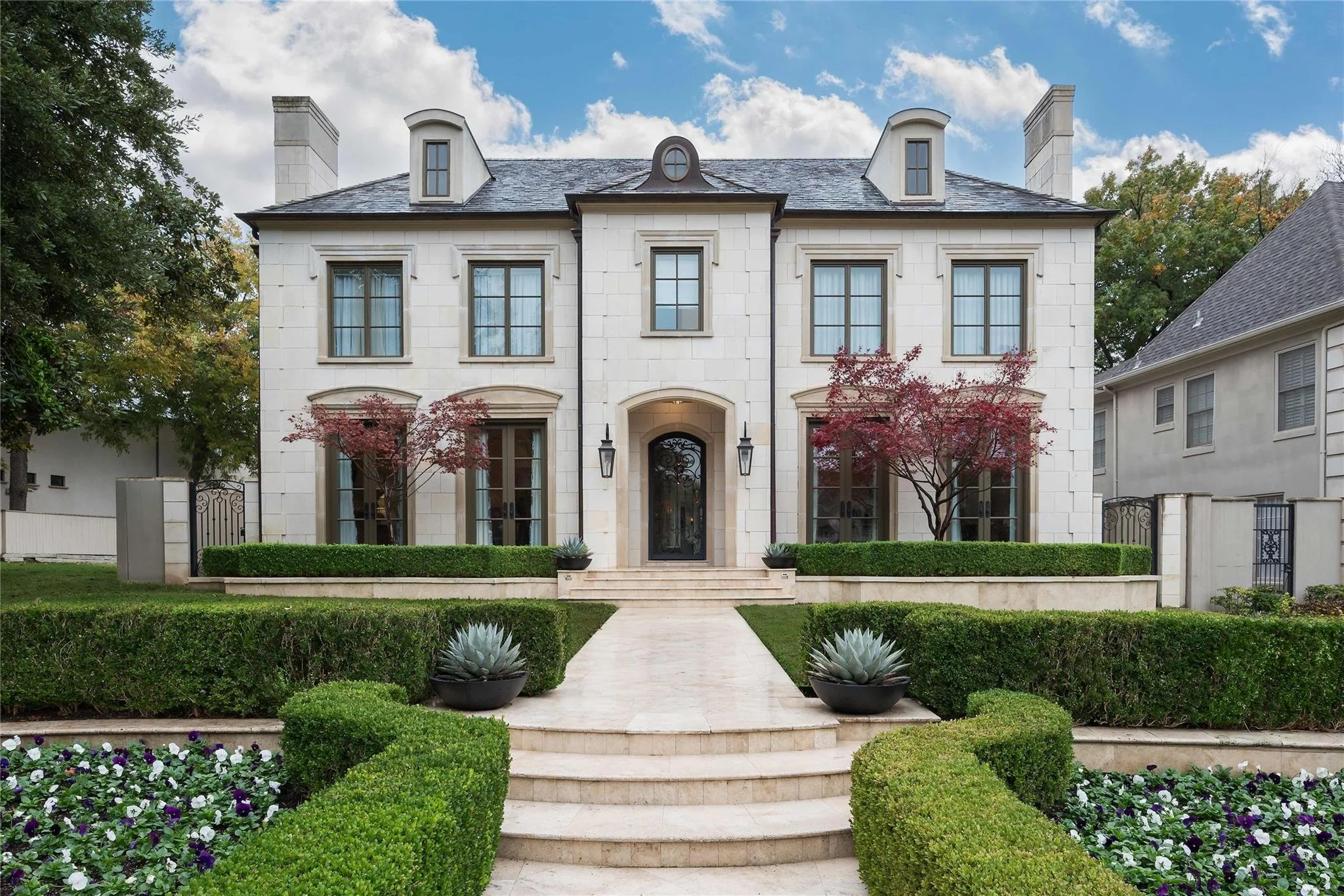 Front view of a large, elegant French Provincial style house with a symmetrical facade, stone exterior, dormer windows, black decorative iron door. Landscaping with trimmed hedges, flowerbeds, Japanese Maples in Old Original Highland Park, Texas.
