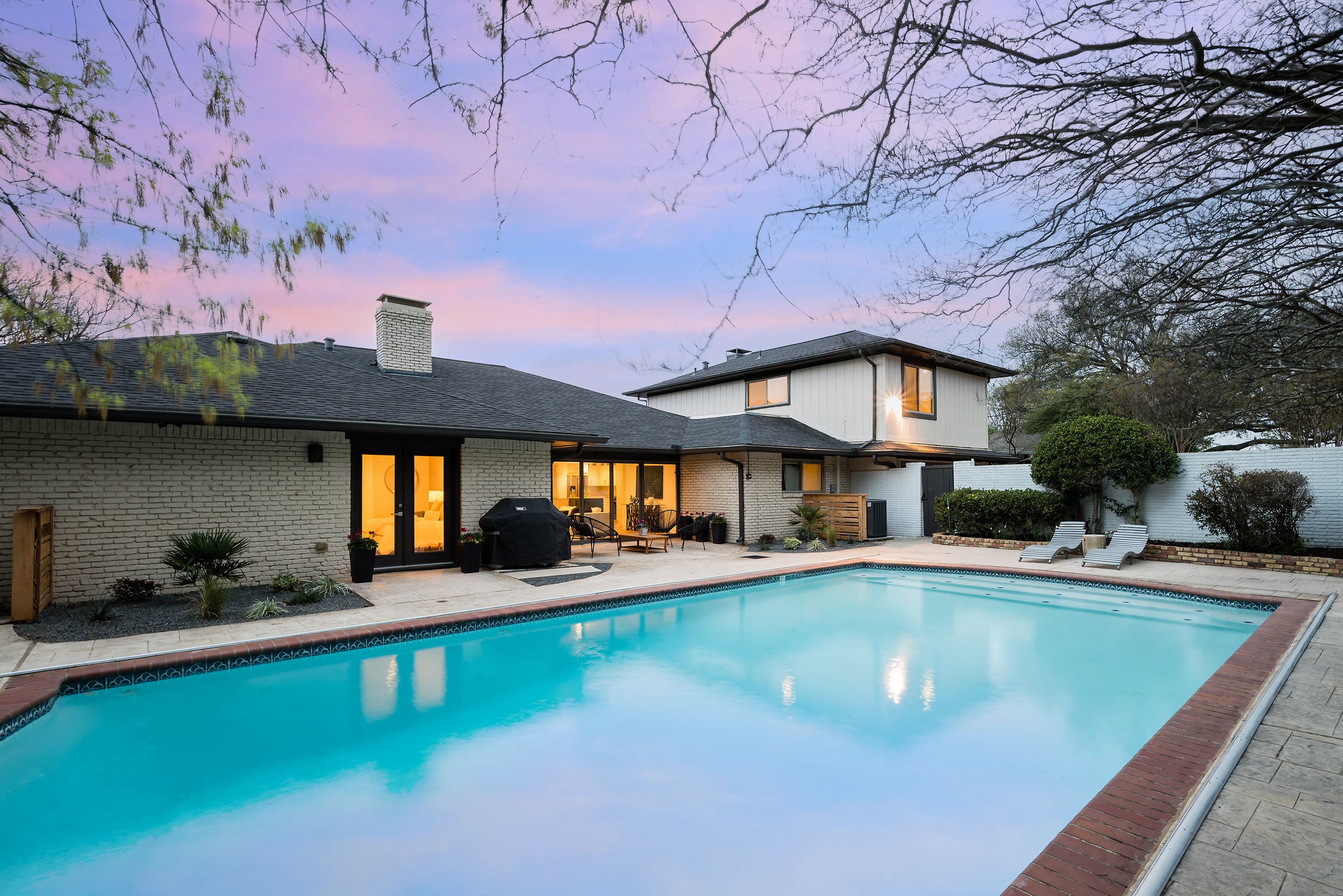 Large backyard with a swimming pool, outdoor furniture, and a two-story house with a chimney at dusk on Mimosa Lane in the Meadows Lane neighborhood of Dallas, TX .