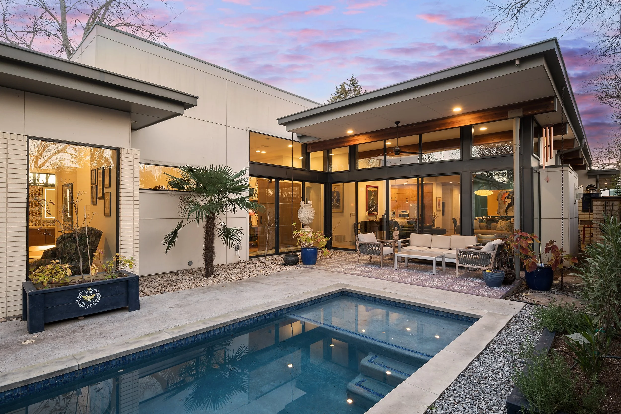 Single story modern house in Little Forest Hills neighborhood of Dallas, Texas with floor to ceiling glass walls, outdoor seating area, and small pool at sunset.