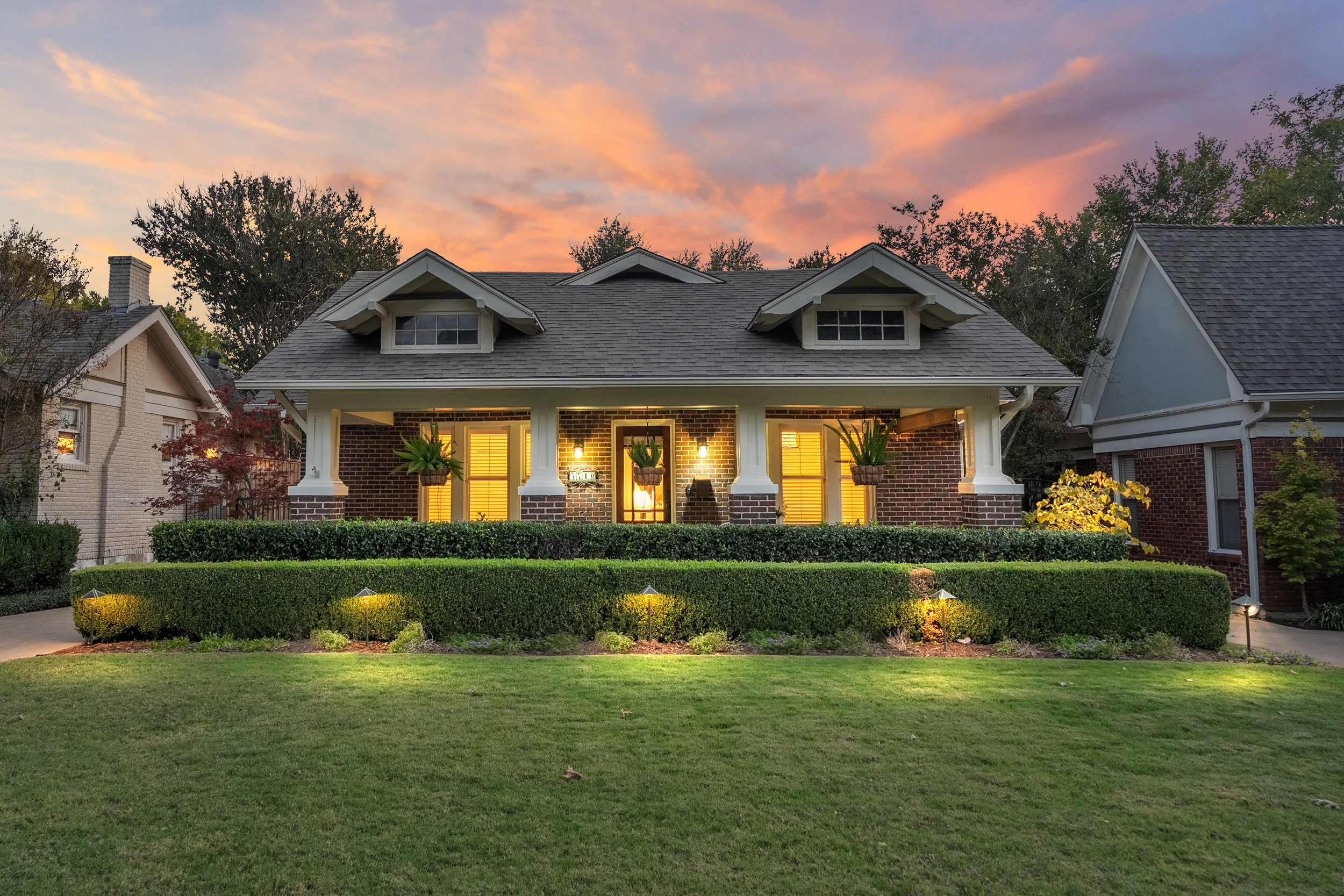 A brick Craftsman early American style home with Prairie style details, landscaped front yard with outdoor lighting at sunset, showing dormer windows, and a front porch with hanging plants located in the M Streets East neighborhood of Dallas, TX.