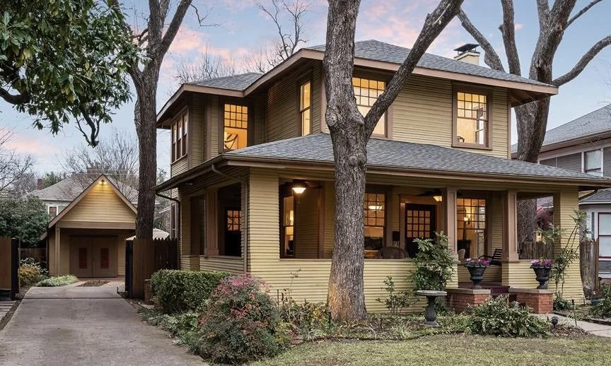 A two-story yellow Prairie style house with large windows and a front porch, surrounded by trees and shrubs, with a driveway leading to a detached garage in the Munger Place neighborhood of Dallas, TX.