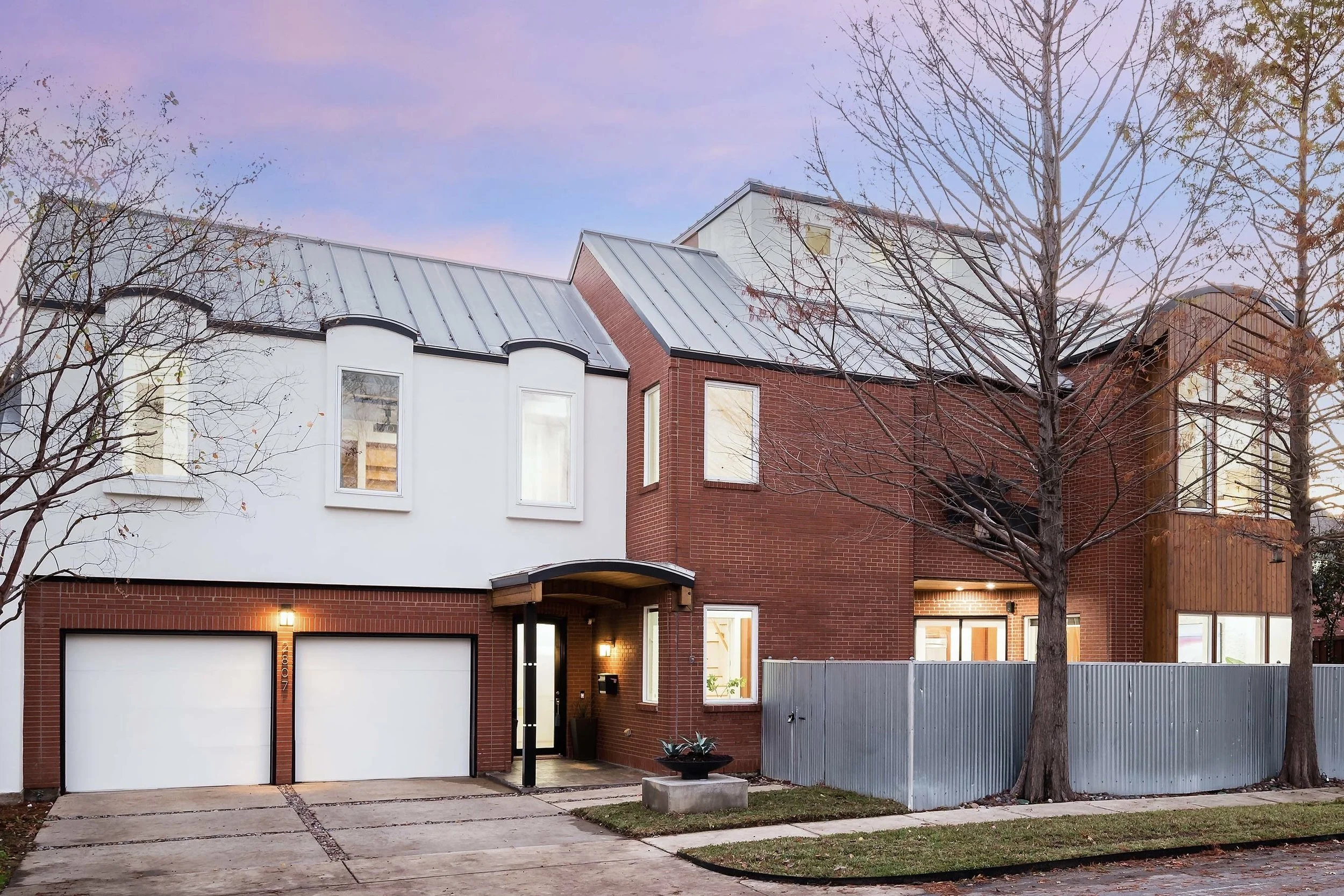Modern residential half duplex with a red brick and white stucco exterior, two garage doors, and pine trees in the late winter lining the front yard during sunset in the Cochran Heights neighborhood of Dallas, TX.