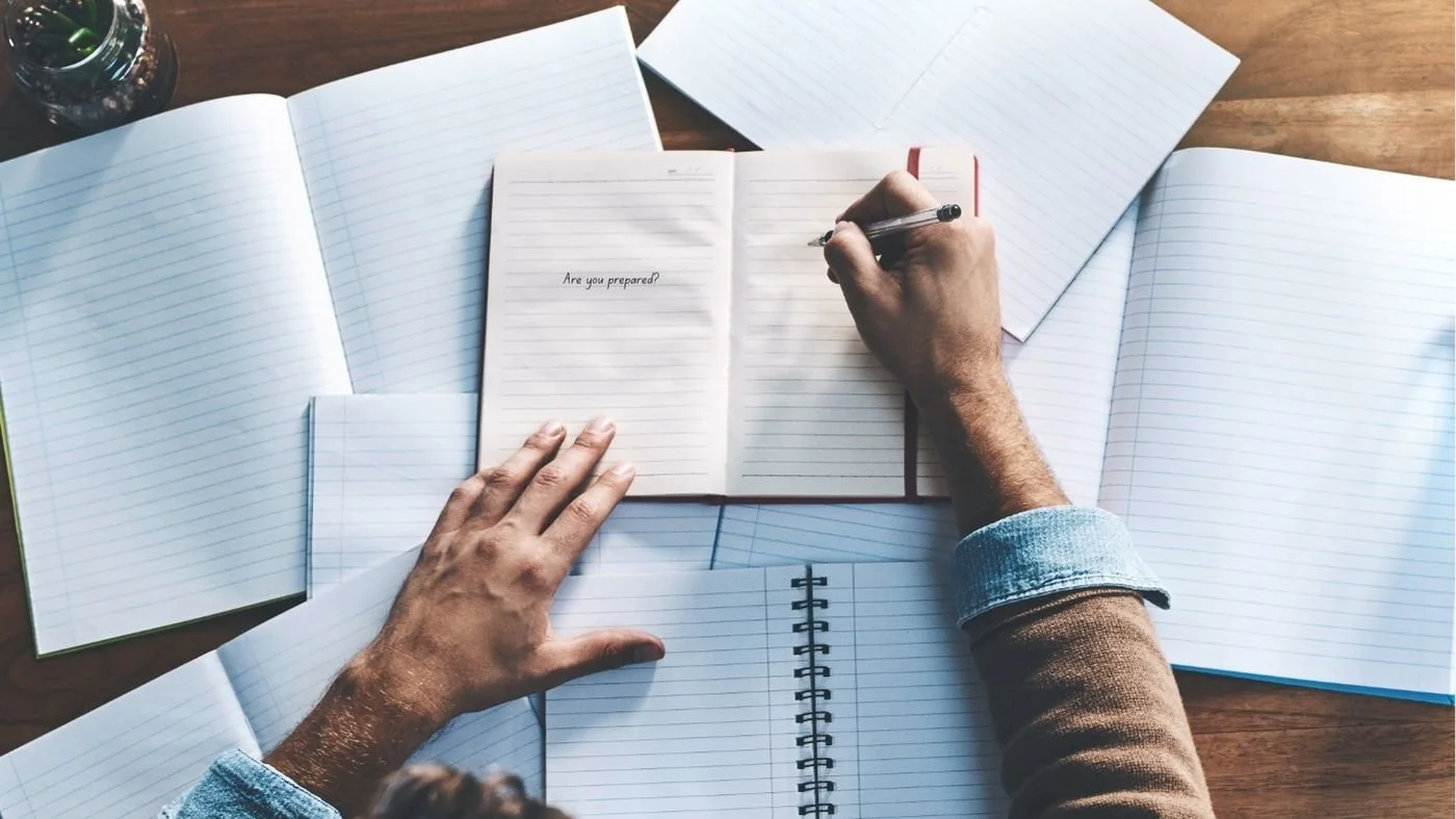 A top down view of a messy desk and a person writing on a notebook in pen that asks "are you prepared?"