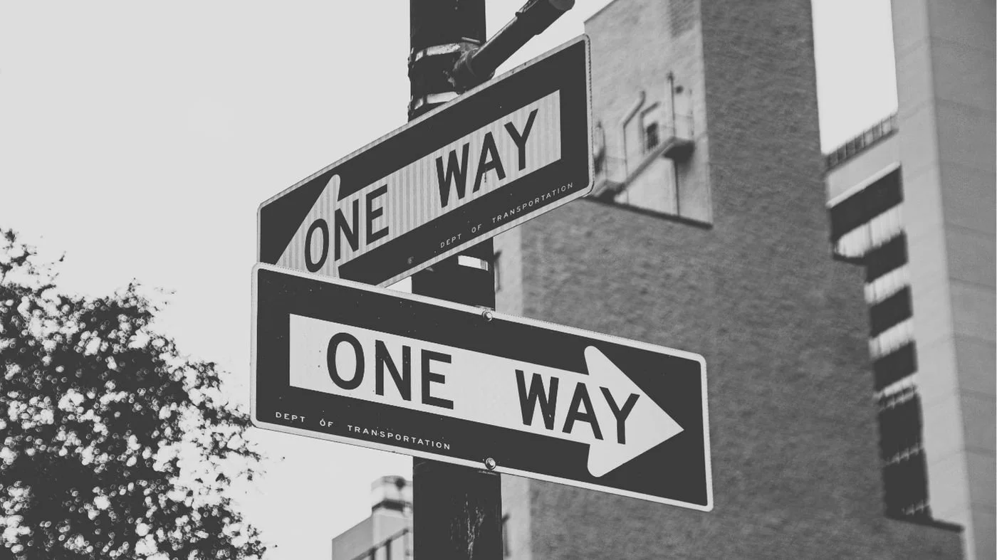 A black and white photo of a street sign pointing in two different directions, creating confusion.