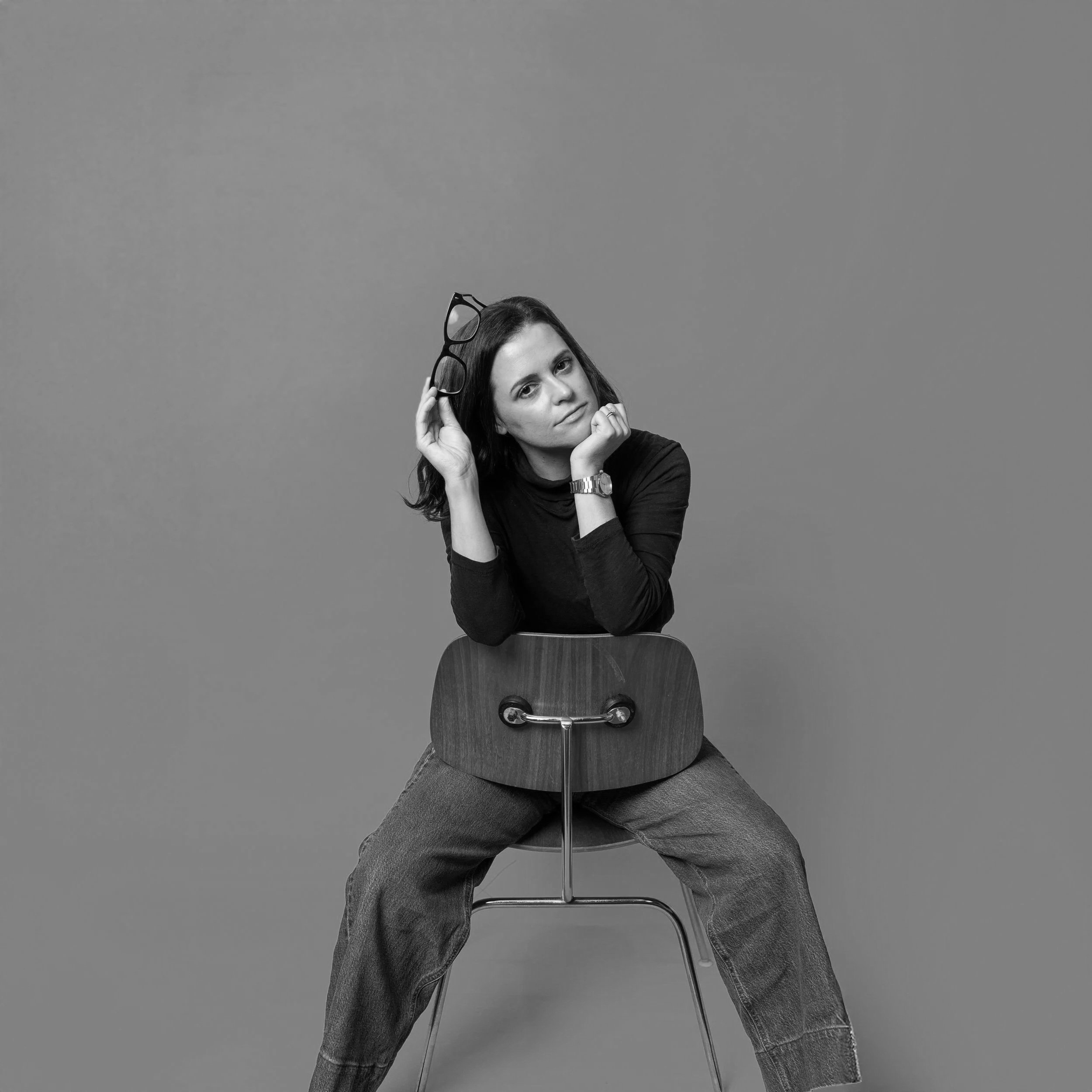 Black-and-white photo of a woman sitting on a chair, holding glasses in her right hand, with her left hand resting on her chin, wearing a long-sleeve shirt and jeans, against a plain background.