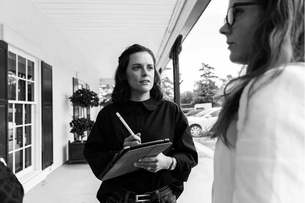 Two women having a conversation outdoors on a porch with a woman holding a tablet and pen, and a woman with glasses facing her, in black and white.
