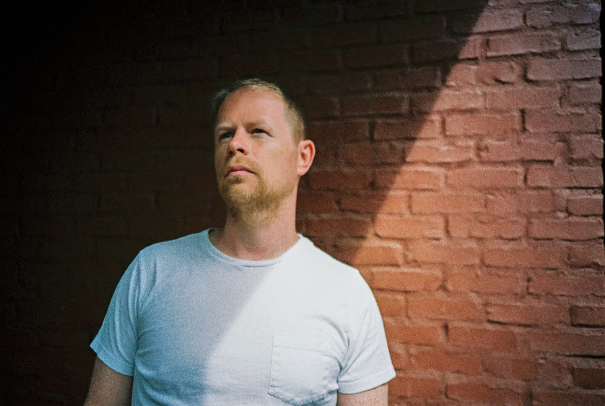A man with short blond hair and a beard, wearing a white t-shirt, standing in partial sunlight against a red brick wall, looking thoughtfully into the distance.