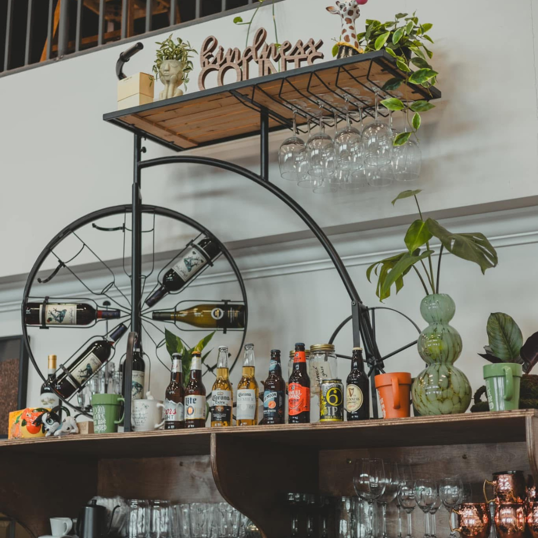 Decorative bar shelf with wine bottles, beer bottles, and glassware, including hanging wine glasses, with plants and a whimsical giraffe figurine on top.