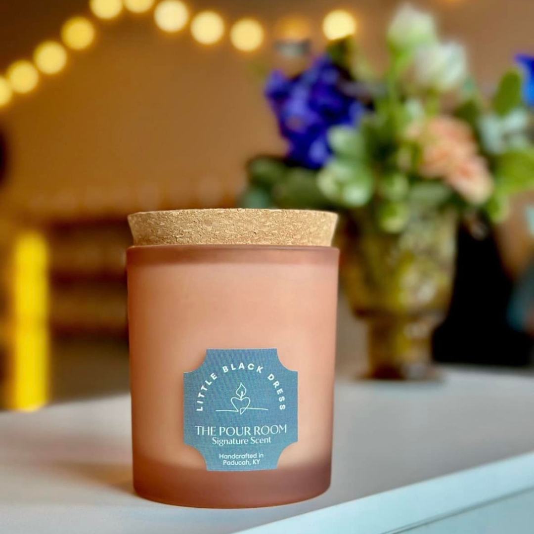 A pink candle with a wooden lid and a blue label that reads 'Little Black Dress' and 'The Pour Room, Signature Scent, Handcrafted in Paducah, KY'. The candle is placed on a white surface with a blurred background featuring flowers in a vase and string lights.