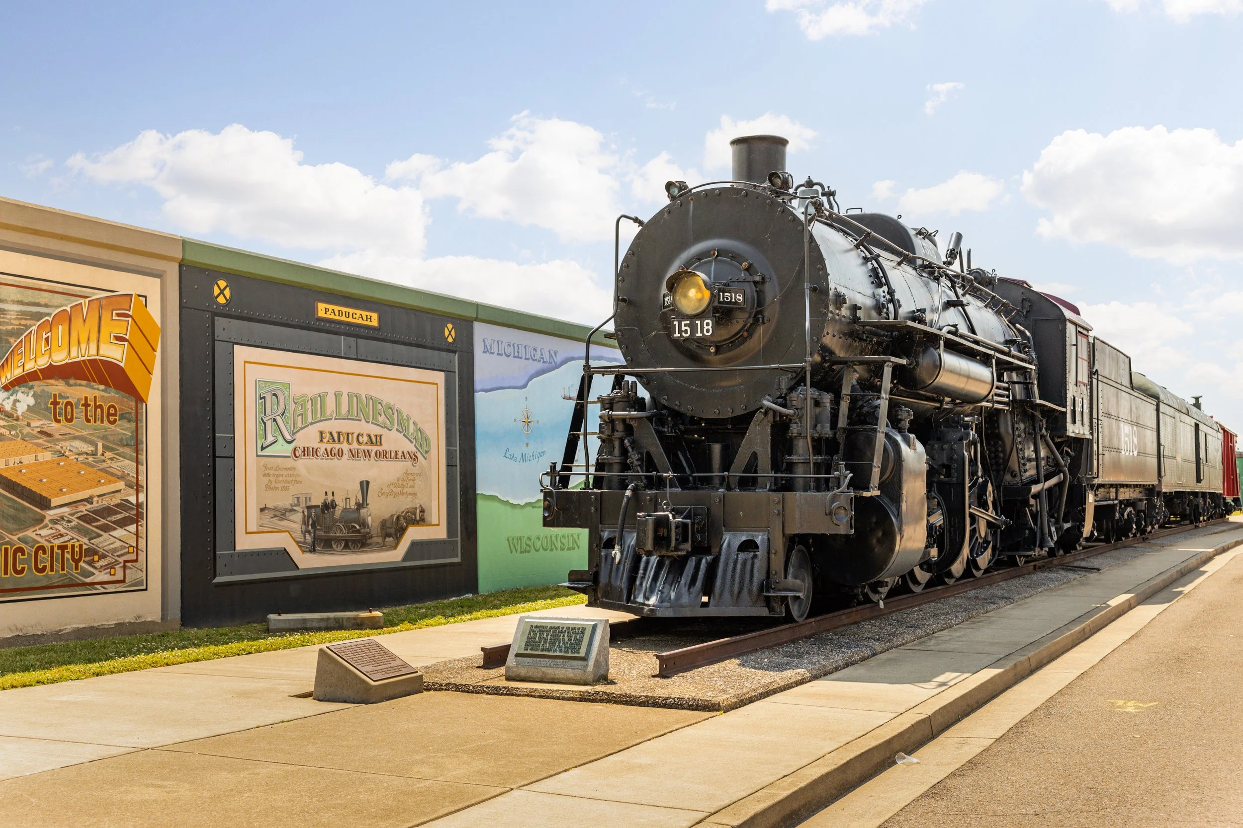 Historical black steam locomotive on display at a museum, with colorful mural and informational plaque nearby.