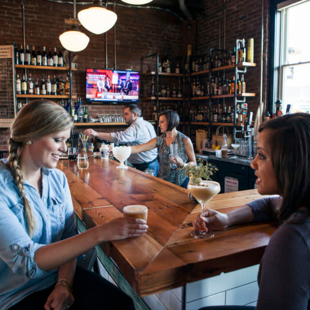 People enjoying drinks at a bar in a cozy, rustic restaurant with brick walls and shelves of liquor bottles.