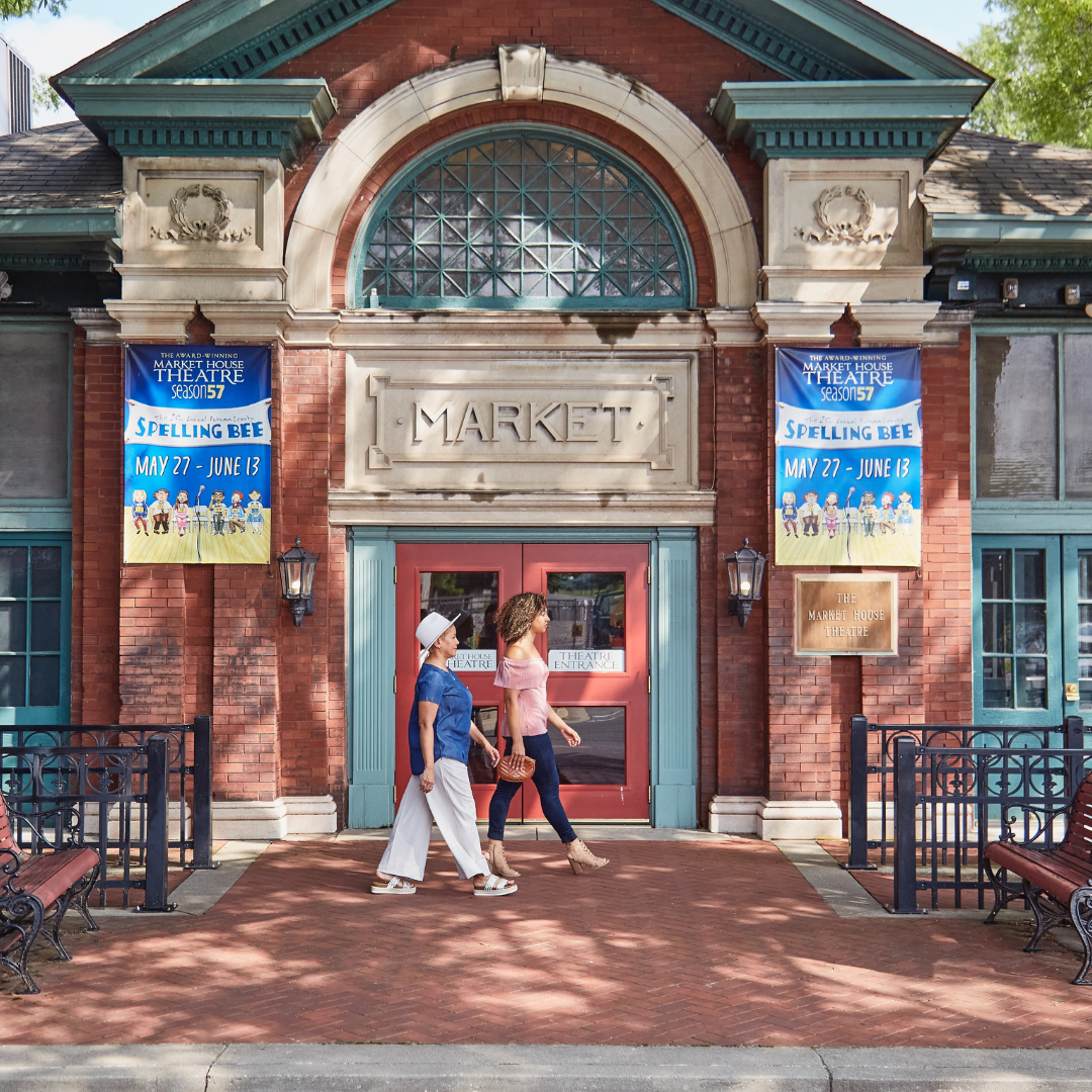 Two women walking in front of the Market House Theatre, a historic brick building with blue and cream accents. Banners on the building promote a play called 'Spelling Bee' running from May 27 to June 13.