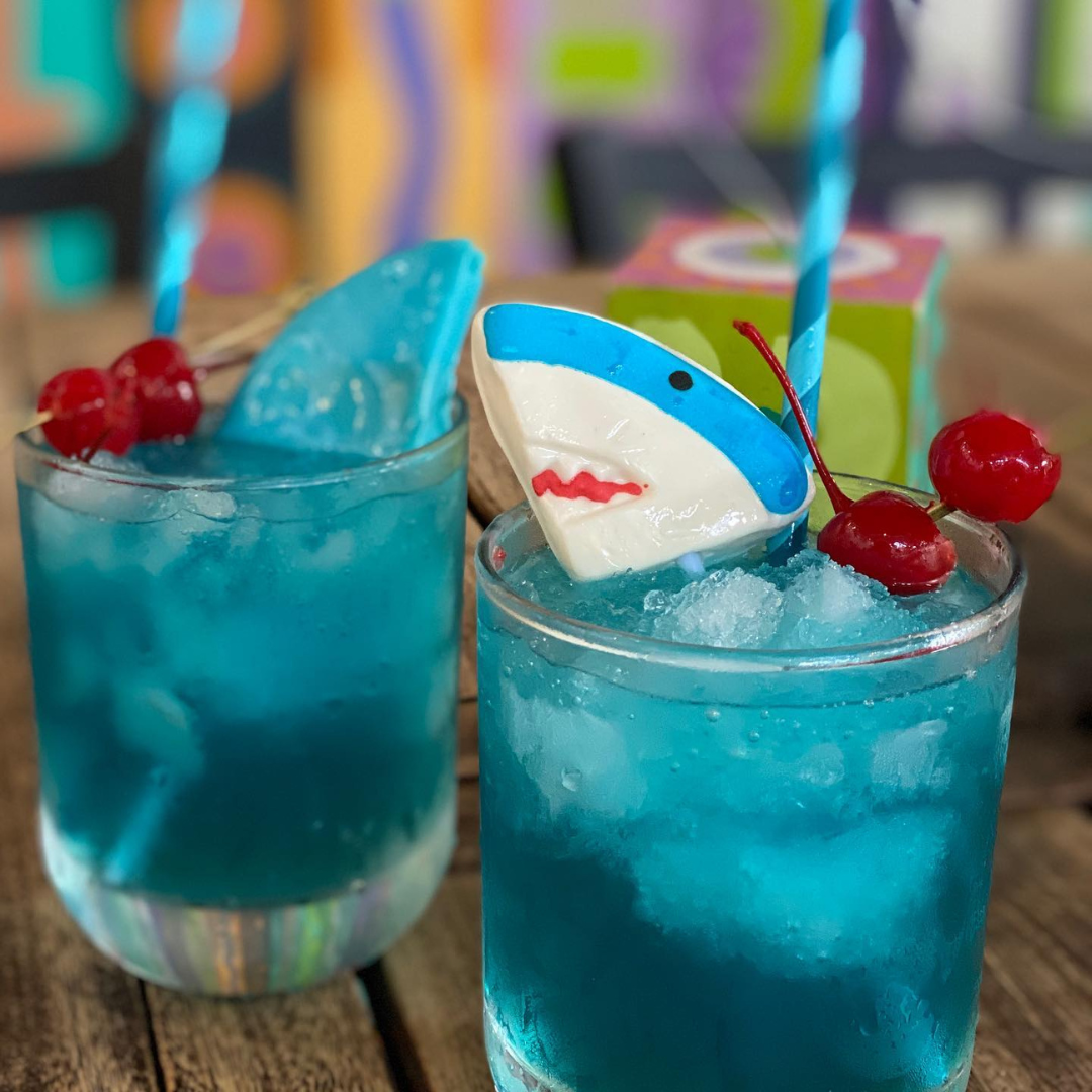 Two blue cocktails with crushed ice, garnished with cherries, a slice of blue and white shark-shaped decoration, and blue striped straws on a wooden table.