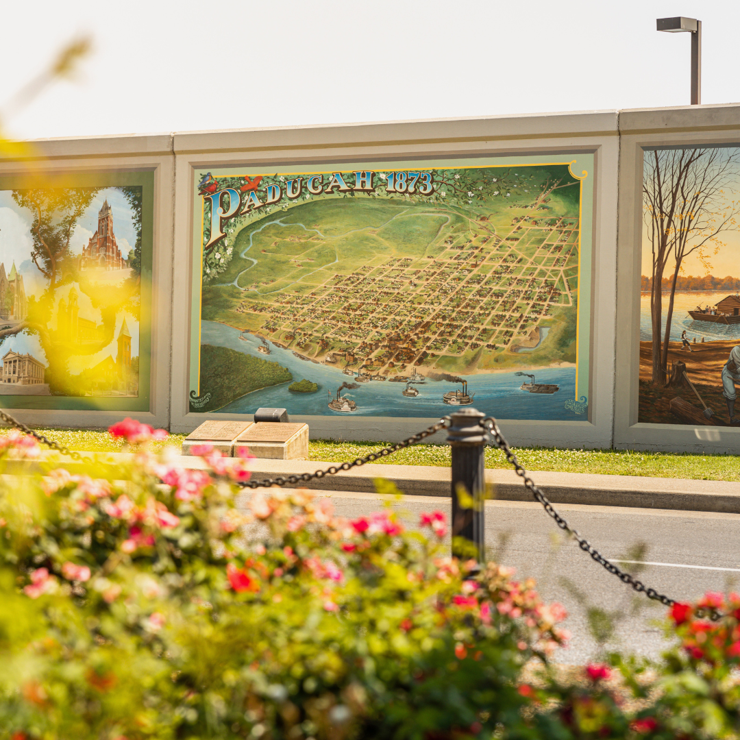 A large illustration of a city map labeled Paduach 1873, with streets, buildings, and a river, displayed on an outdoor wall with a chain fence and pink flowers in the foreground.