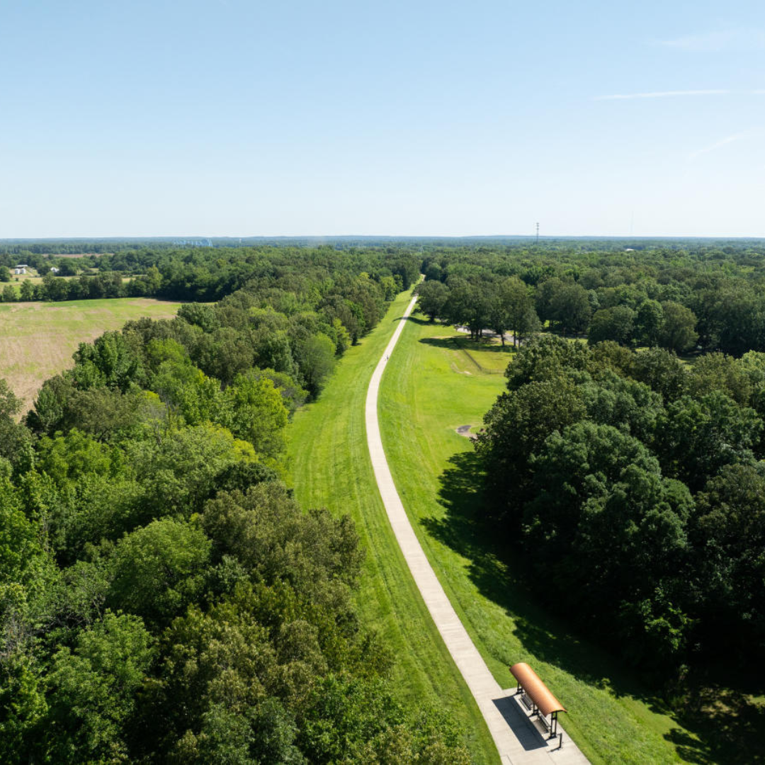 A scenic aerial view of a winding pathway through lush green trees and grass on a bright, sunny day.