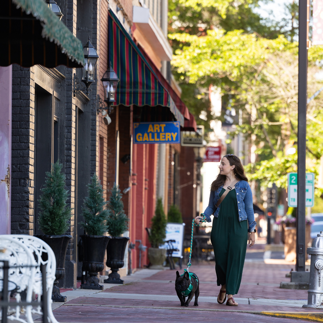 A woman in a green dress and denim jacket walks her black dog on a leash down a sidewalk lined with small potted evergreen trees. She is looking to her left and smiling. Buildings with colorful awnings and a sign for an art gallery are visible in the background, as well as trees and street signs.