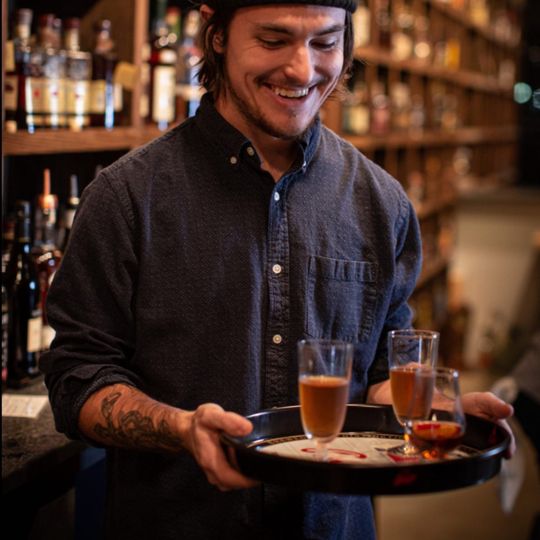 A smiling man with a tattoo on his left arm carrying a tray with three glasses of amber-colored drinks inside a bar or restaurant.