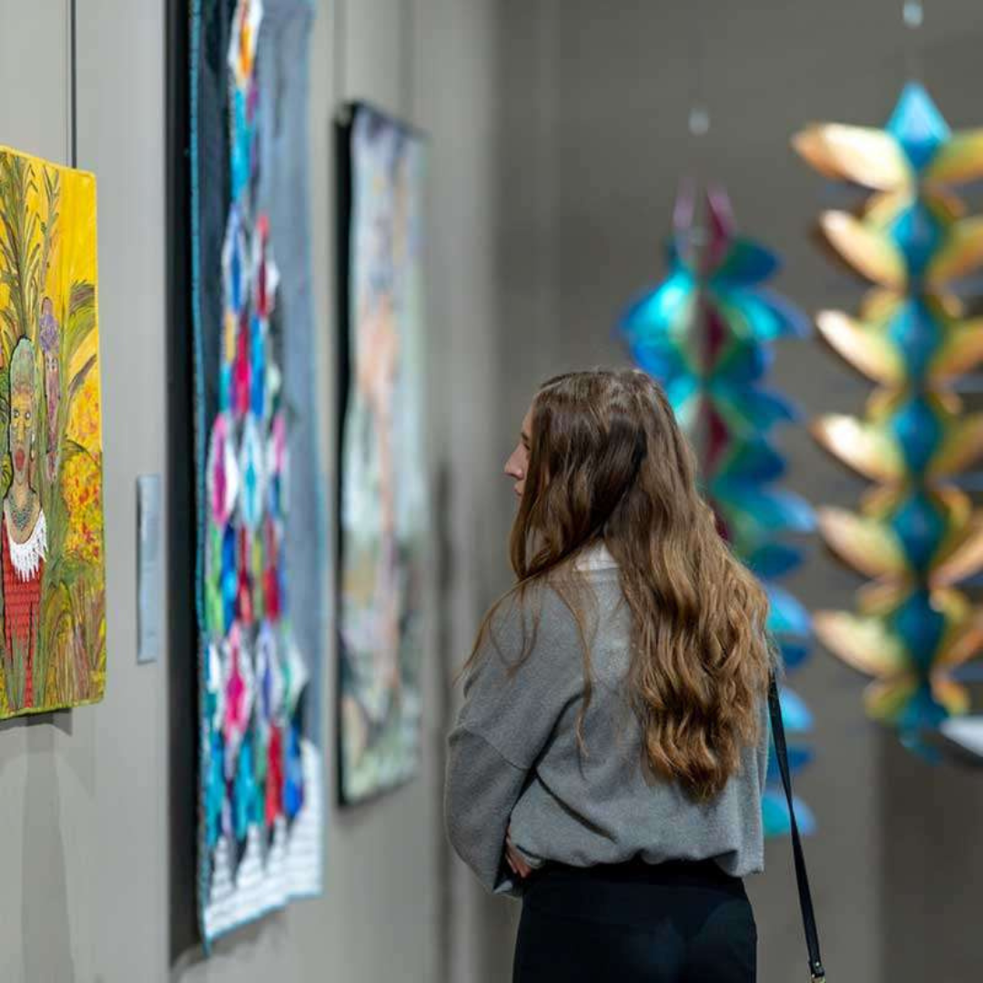 A young woman with long wavy brown hair observes artwork on a gallery wall, dressed in a gray top and black pants.