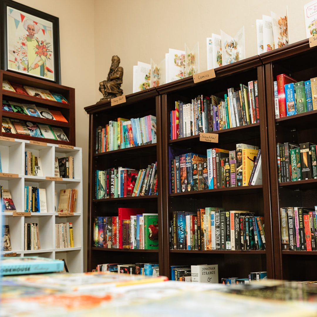 Bookshelves filled with various books and comics, with labels on the shelves indicating categories like science fiction, fantasy, comics, and young adult. There is a framed artwork and a small statue on top of the shelves.