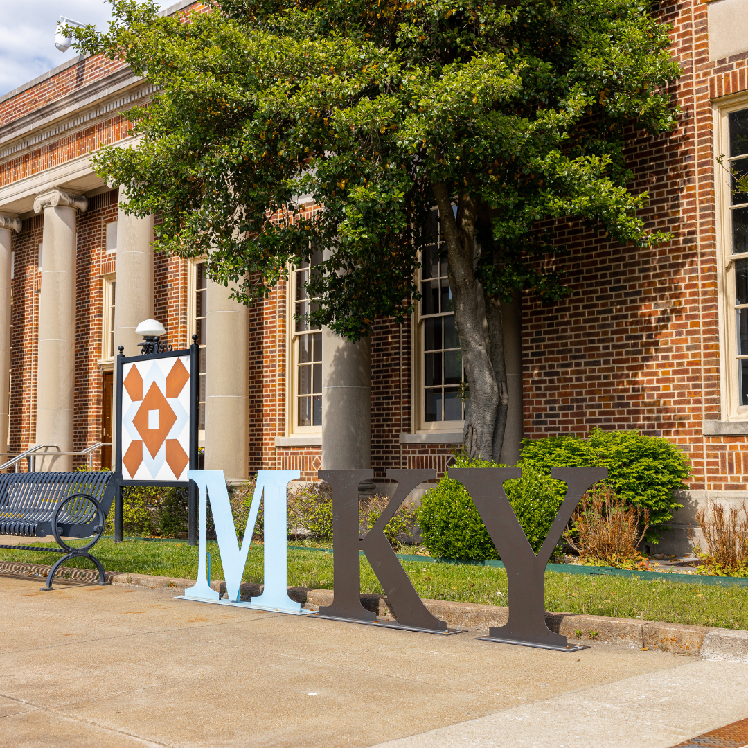 Large colorful letters spelling 'MKY' outside a brick building with windows, trees, and greenery.