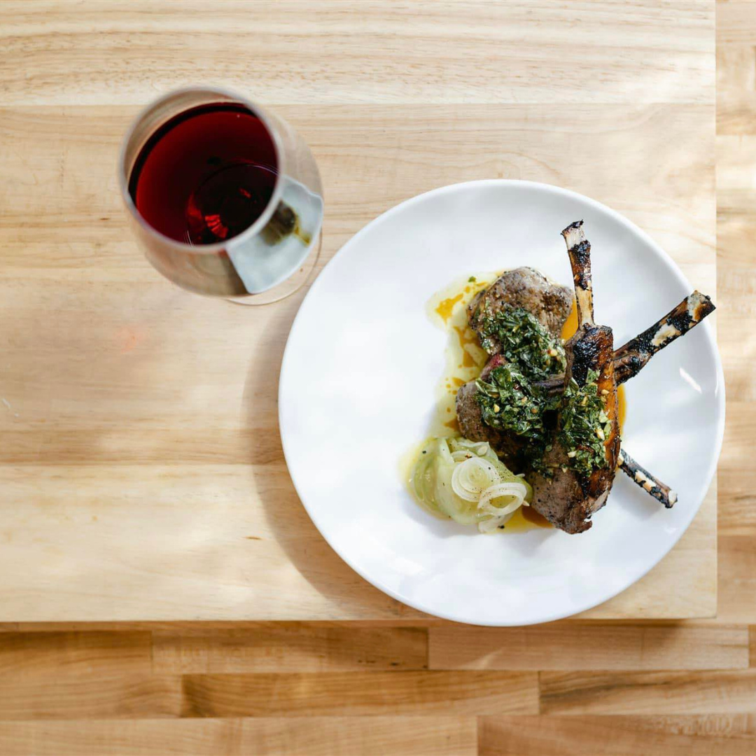 A white plate with grilled lamb chops topped with chimichurri sauce, served with sliced onions and a glass of red wine on a wooden table.