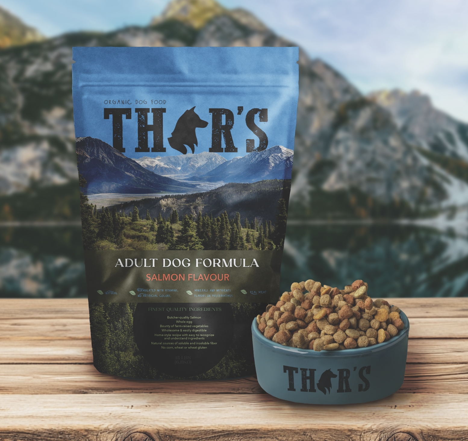 A bag of Thors organic dog food with salmon flavor and a bowl filled with kibble, set on a wooden surface outdoors against a backdrop of mountains and a lake.