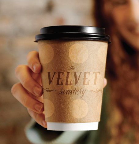 Person holding a paper coffee cup with a black lid, labeled 'The Velvet Roastery'