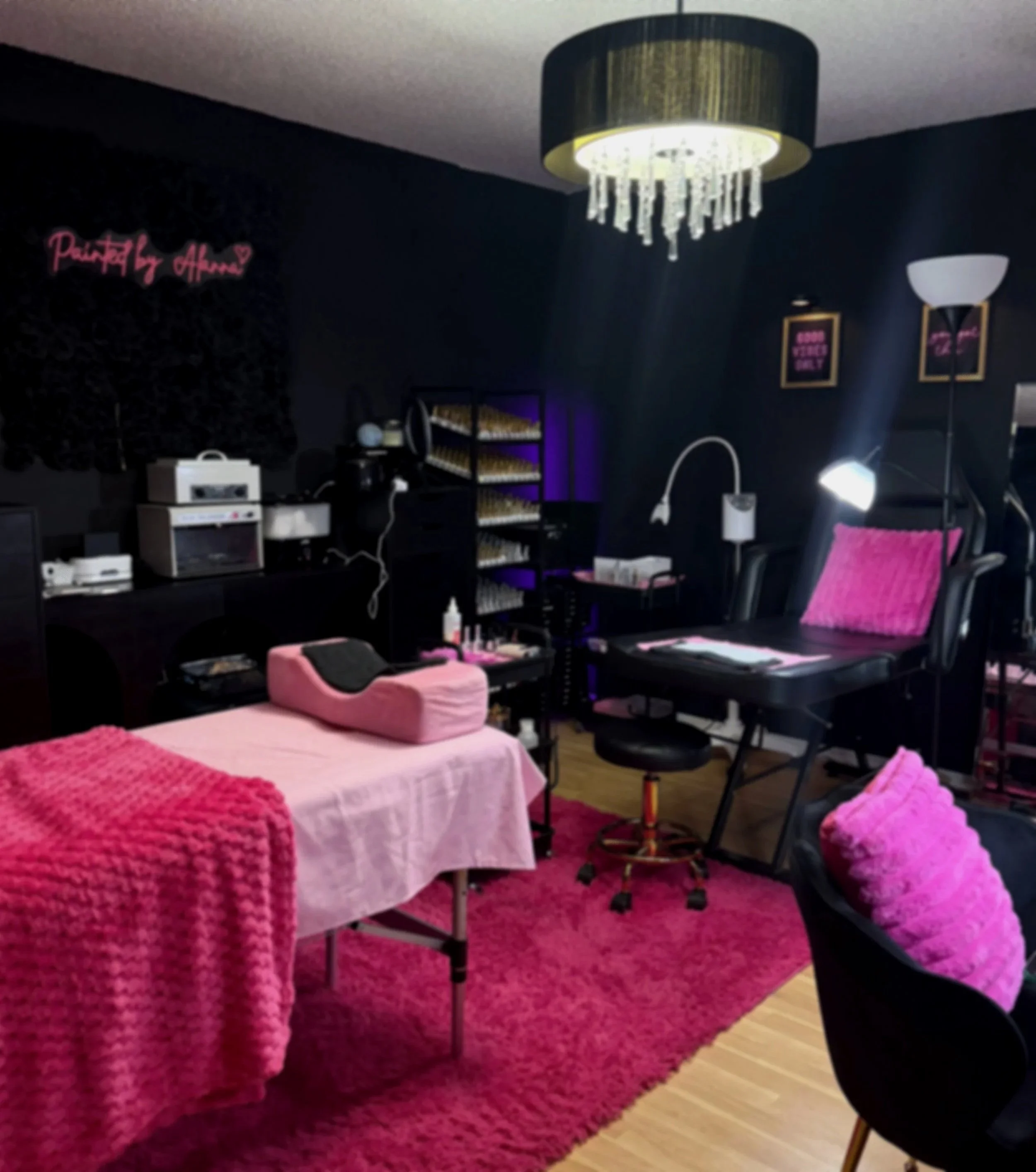 A cozy nail salon or beauty treatment room with black walls and pink accents, including a pink fuzzy blanket and pillows, a massage or treatment table, and various salon equipment and lighting.