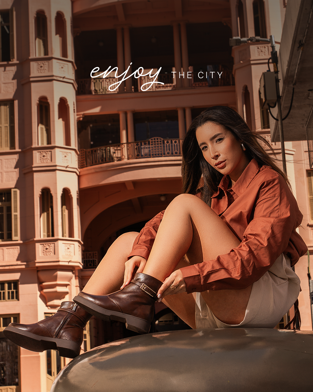 Mulher sentada em um objeto metálico, vestindo uma blusa marrom e saia bege, ao fundo edifícios históricos de cor rosa.