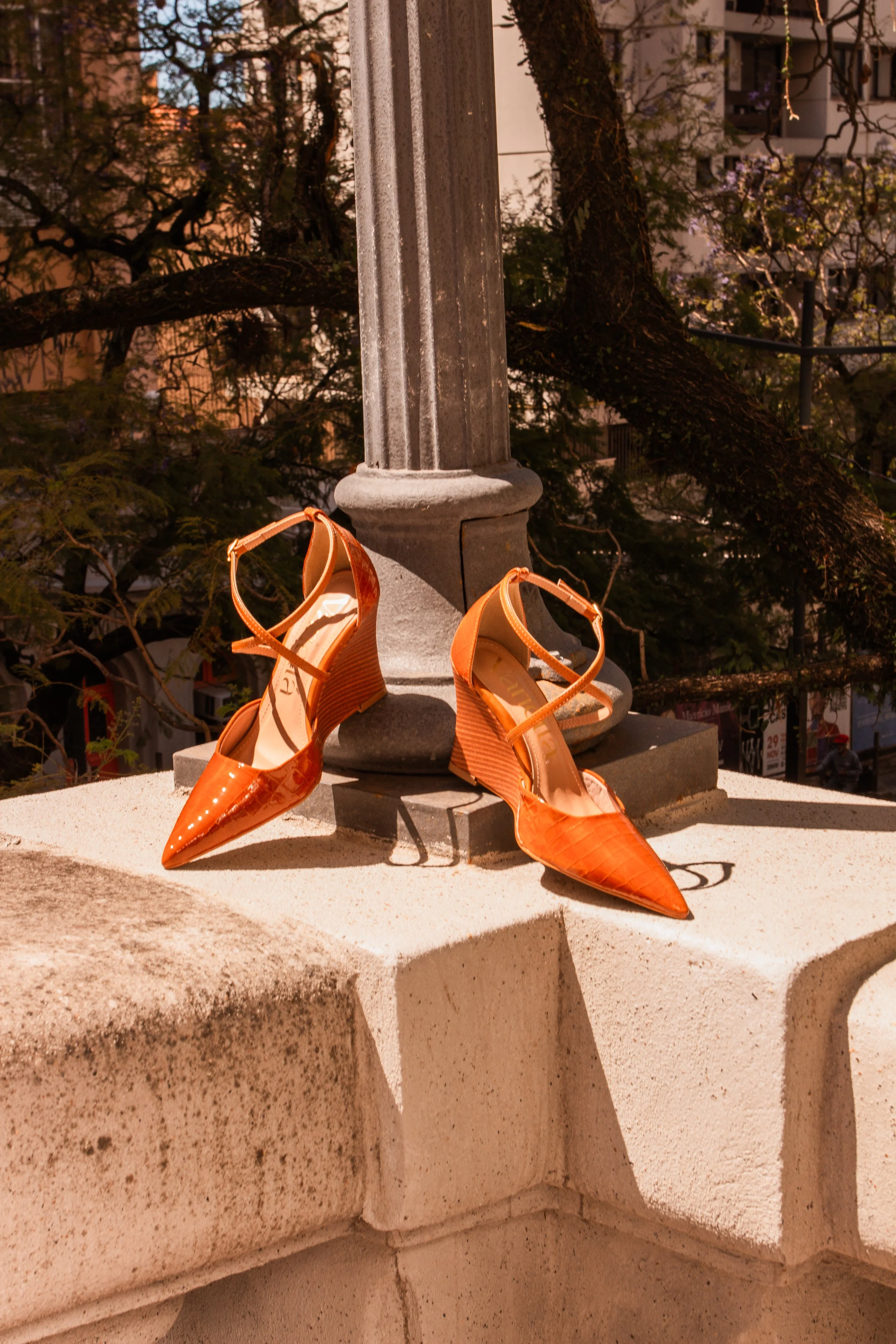 Um par de sapatos de salto alto na cor laranja, posicionado em cima de uma parede de pedra. Ao fundo há uma árvore e edifícios, sugerindo uma cena ao ar livre durante o dia.
