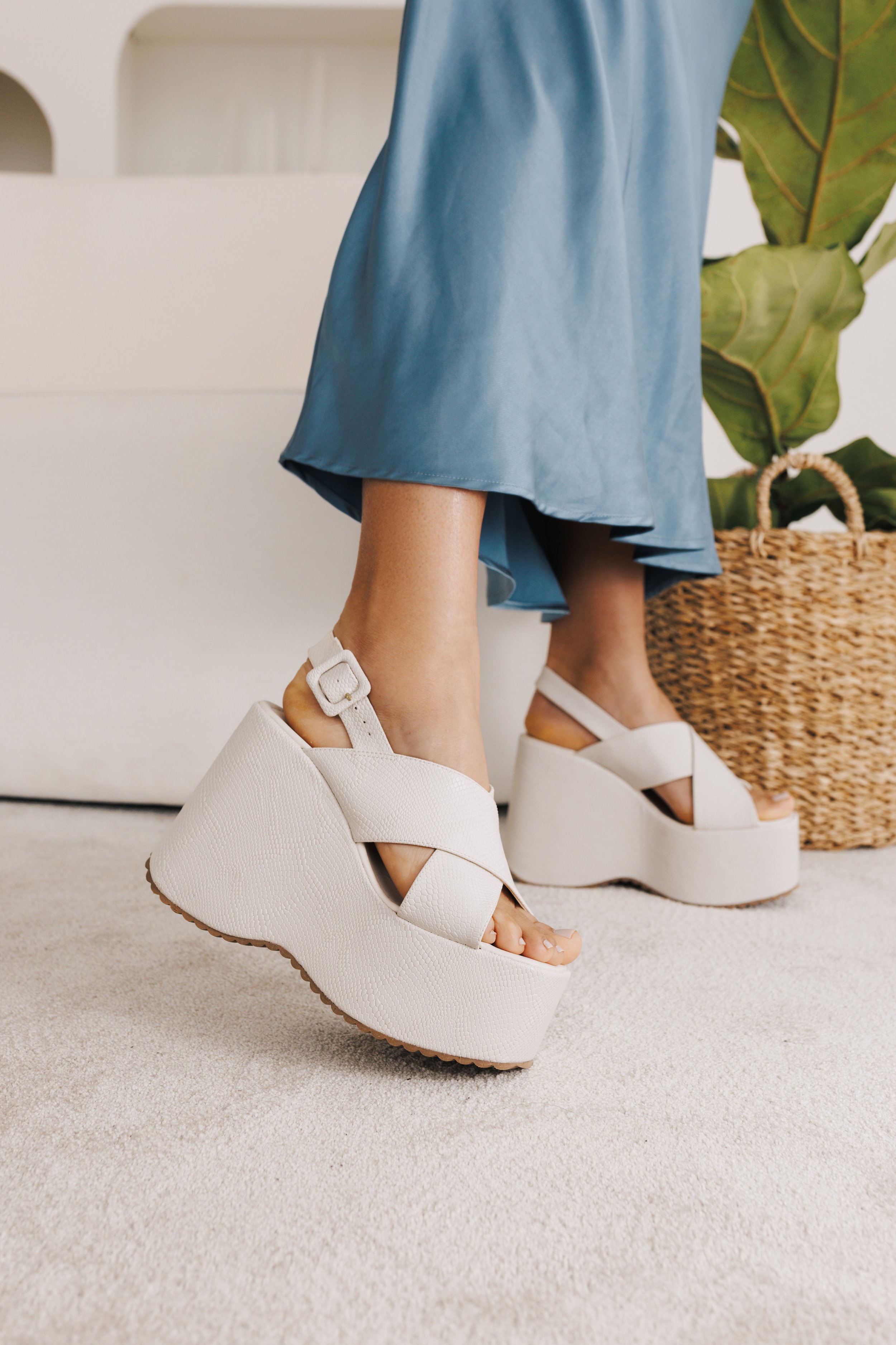 Mulher usando sandálias plataforma brancas com cós largo e fivela, vestindo saia azul e em um ambiente com tapete claro e planta no fundo.