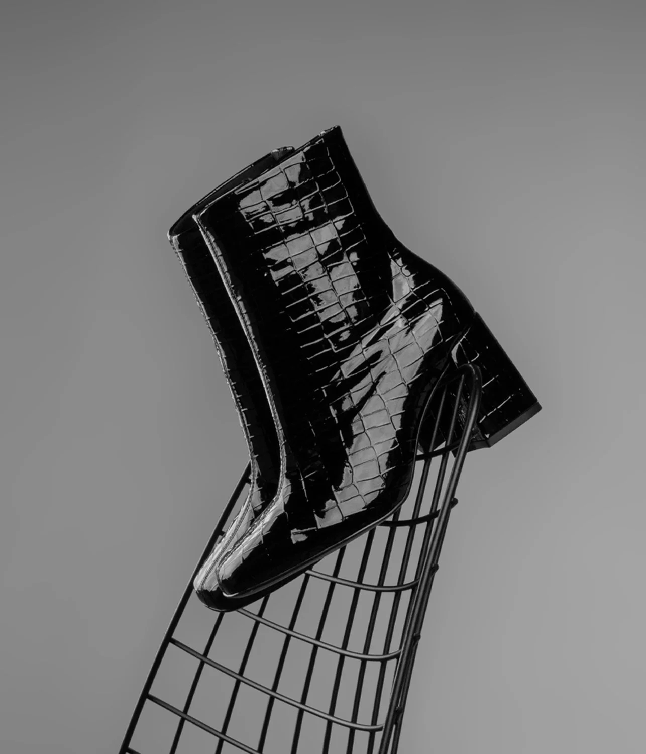 Bota preta de couro com estampa de crocodilo, apoiada em uma estrutura metálica, em fundo cinza.